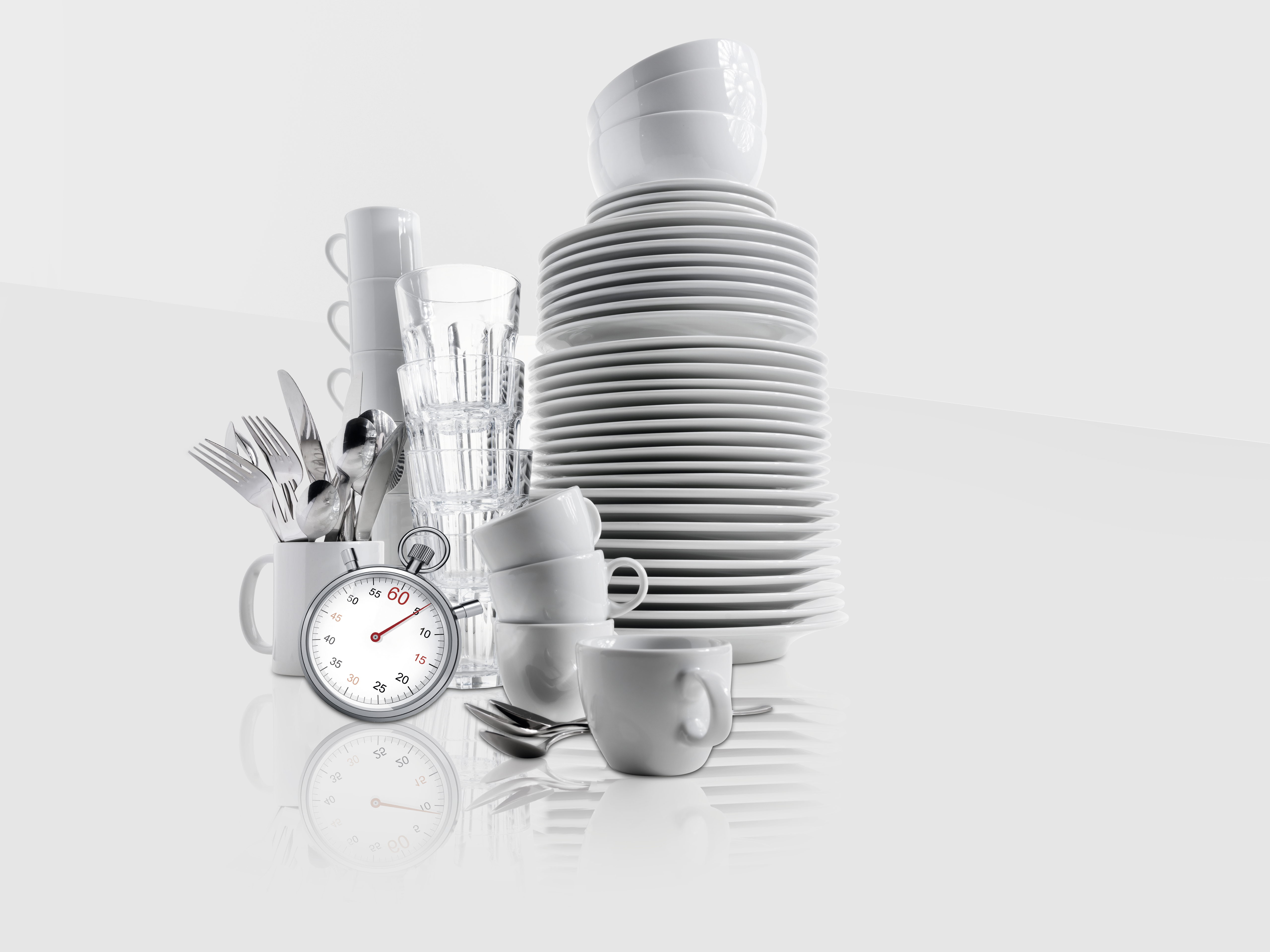 Stack of clean white plates, cups, glasses, and cutlery with a stopwatch on a reflective surface against a light gray background.