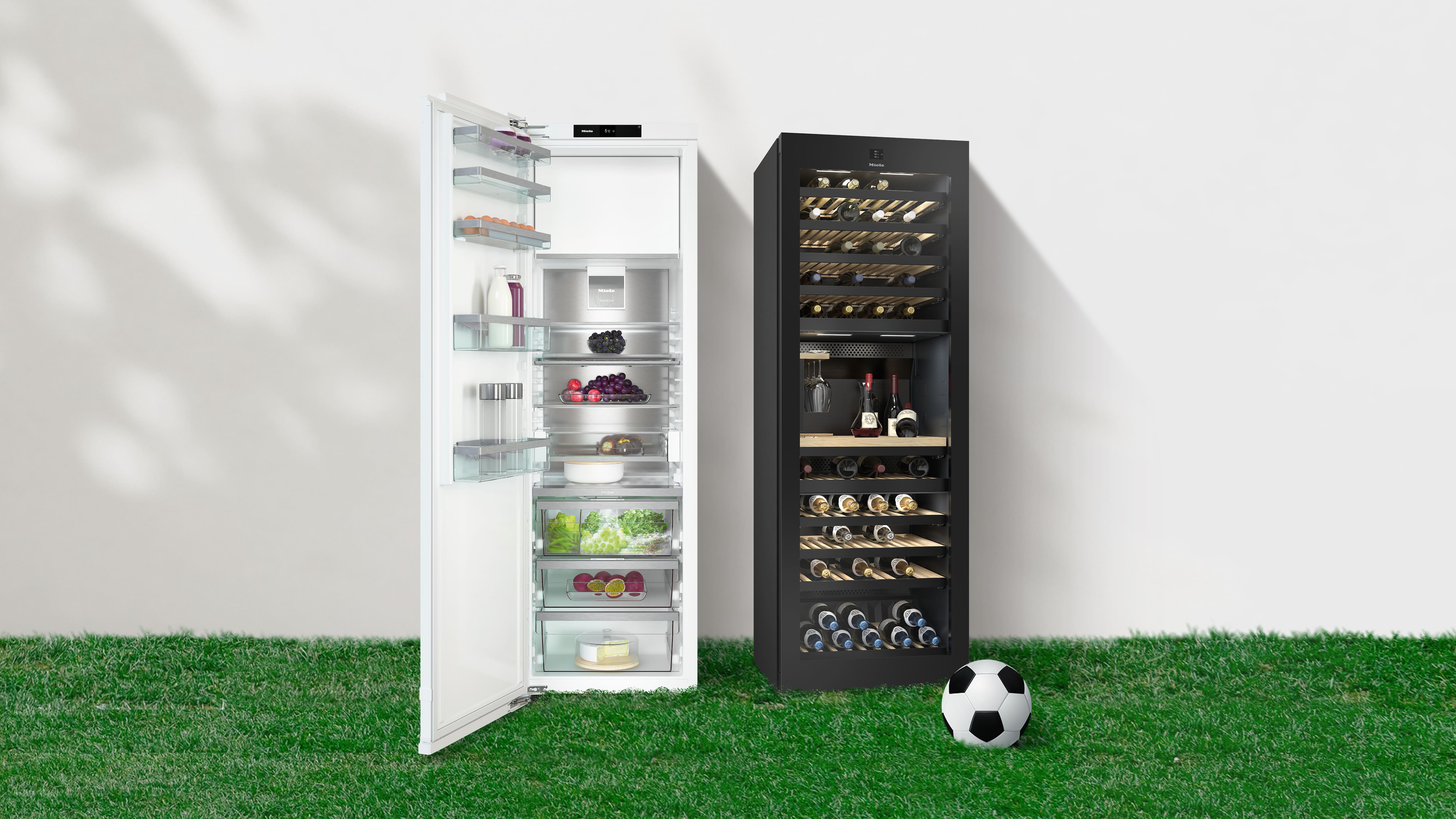 Geöffneter weisser Kühlschrank und schwarzer Weinkühler nebeneinander auf einer Rasenfläche, beide gefüllt mit Lebensmitteln und Flaschen; daneben liegt ein Fussball.