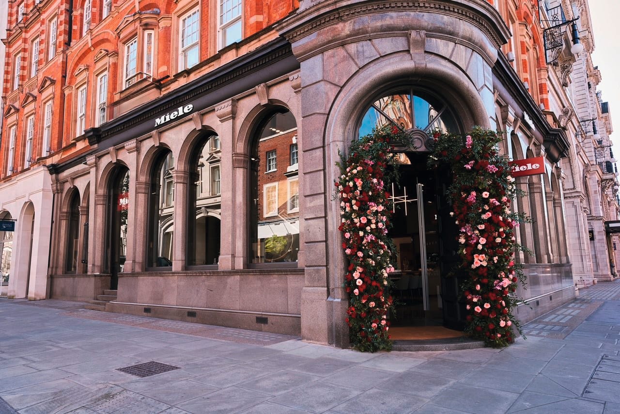 Street view of a historic corner building with arched windows and a Miele storefront; the entrance is framed by a large red and pink floral archway. Street view of a historic corner building with arched windows and a Miele storefront; the entrance is framed by a large red and pink floral archway.