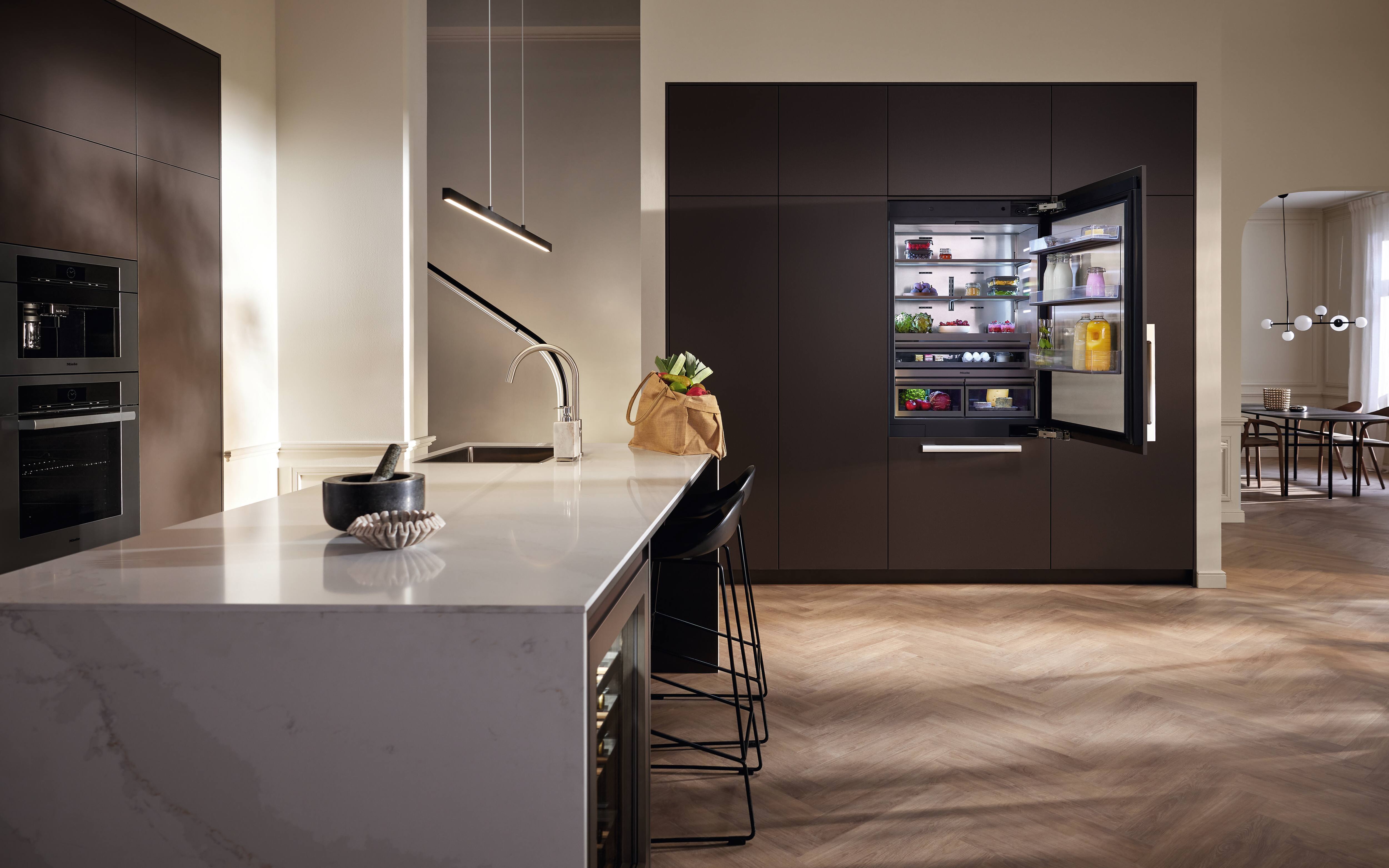Cozinha contemporânea com armários escuros, uma geladeira embutida aberta, uma ilha de mármore com pia e dois bancos pretos, uma sacola de compras sobre a bancada, luminárias pendentes e piso de madeira em padrão espinha de peixe, com vista para a área de jantar. Cozinha contemporânea com armários escuros, uma geladeira embutida aberta, uma ilha de mármore com pia e dois bancos pretos, uma sacola de compras sobre a bancada, luminárias pendentes e piso de madeira em padrão espinha de peixe, com vista para a área de jantar.