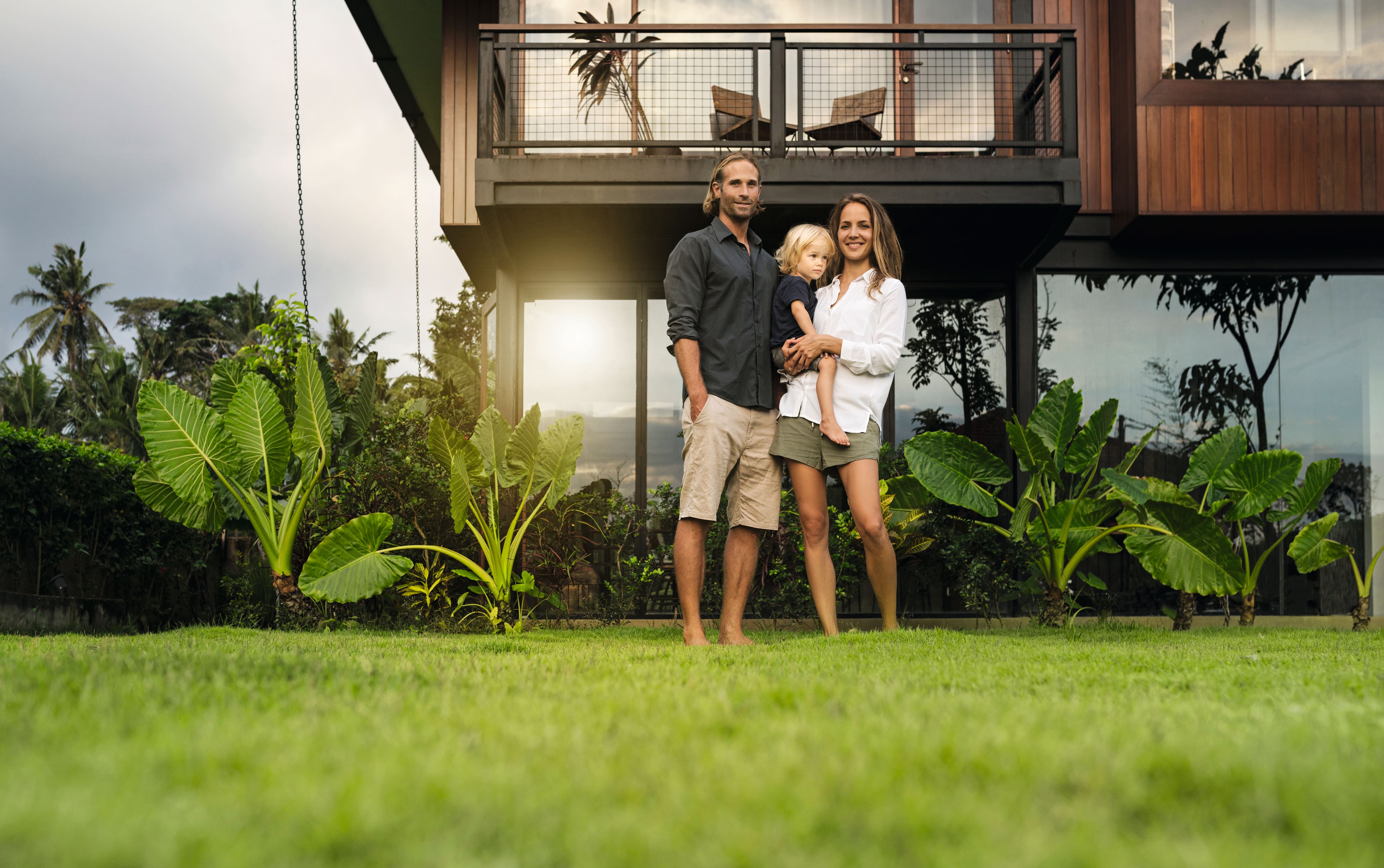 Une famille de trois personnes, pieds nus, debout sur une pelouse devant une maison moderne, avec des plantes tropicales et un reflet du soleil en arrière-plan. Une famille de trois personnes, pieds nus, debout sur une pelouse devant une maison moderne, avec des plantes tropicales et un reflet du soleil en arrière-plan.