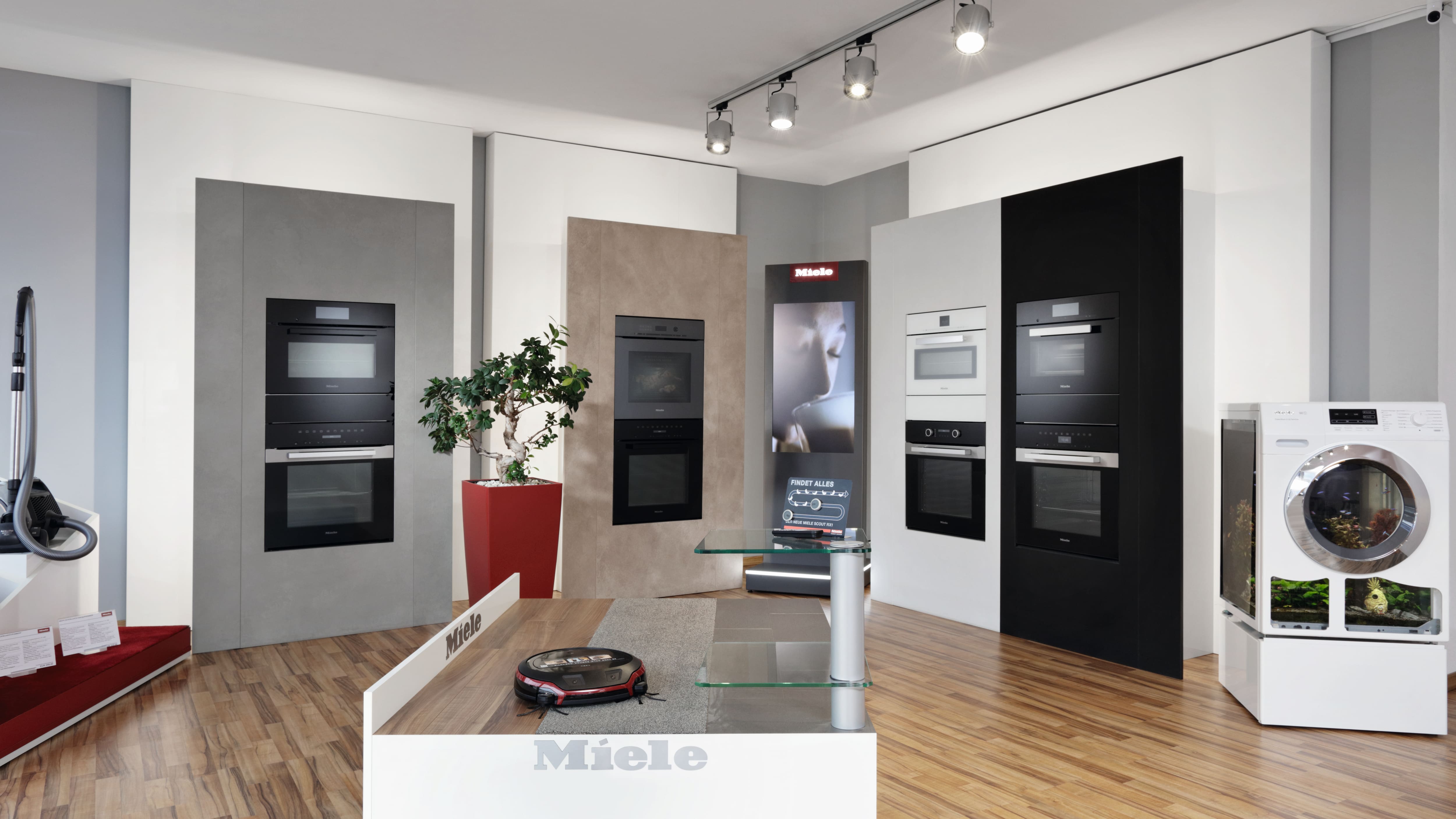 Moderner Geräteschauraum mit Miele-Branding, mehrere eingebaute Öfen in Wanddisplays, eine weiße Waschmaschine rechts, ein Saugroboter auf einer Plattform in der Mitte und Schienenbeleuchtung über Holzböden mit einer Topfpflanze.