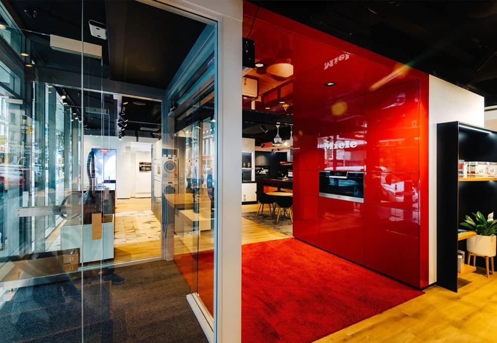 Modern appliance showroom featuring a glossy red Miele display wall, glass partitions, wood floors, and a demo kitchen area.