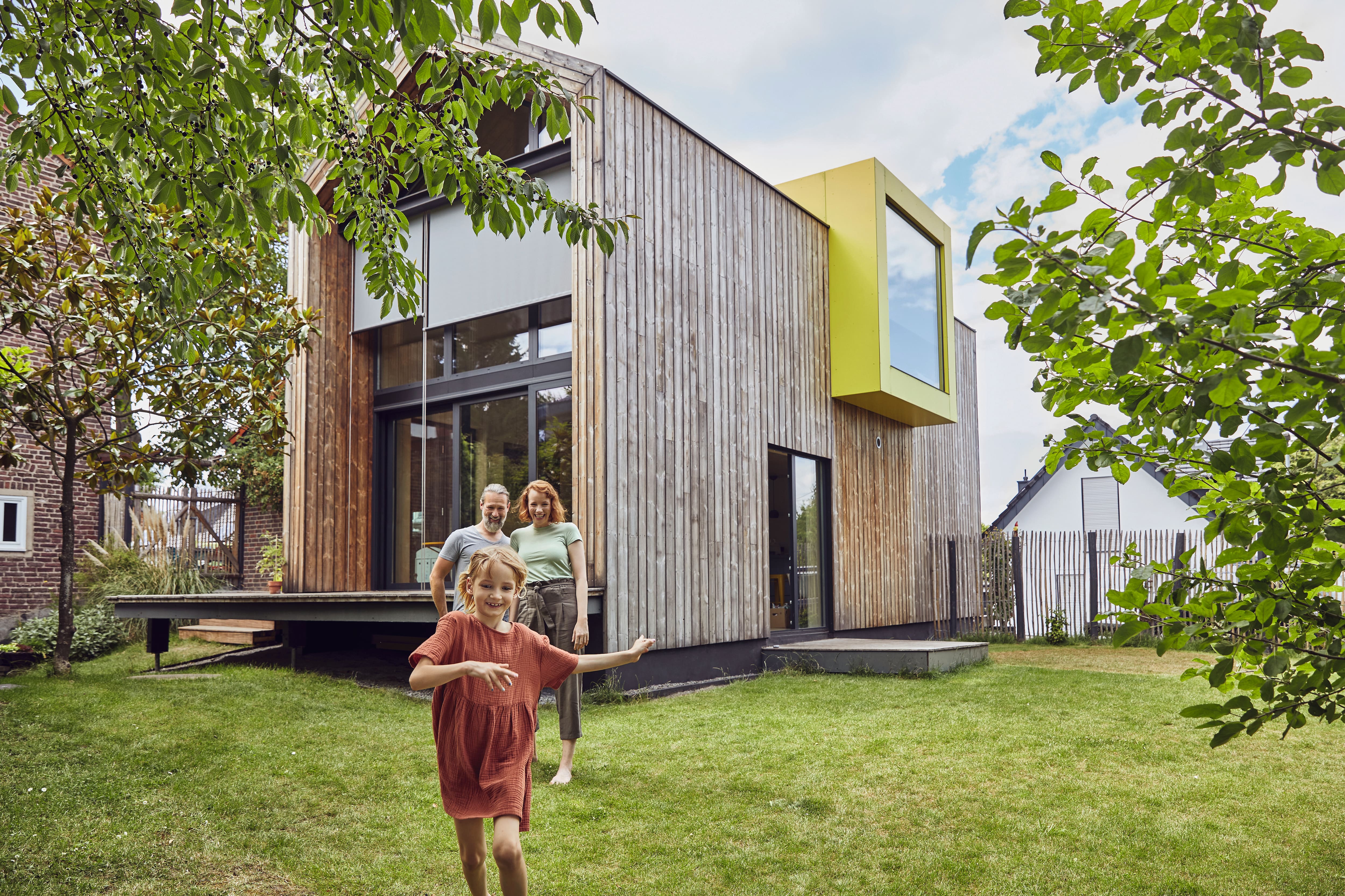 Modernes Holzhaus mit grossen Fensterflächen und einem gelben Erker. Im grünen Garten unter schattenspendenden Bäumen spielen Menschen und geniessen die naturnahe Umgebung.