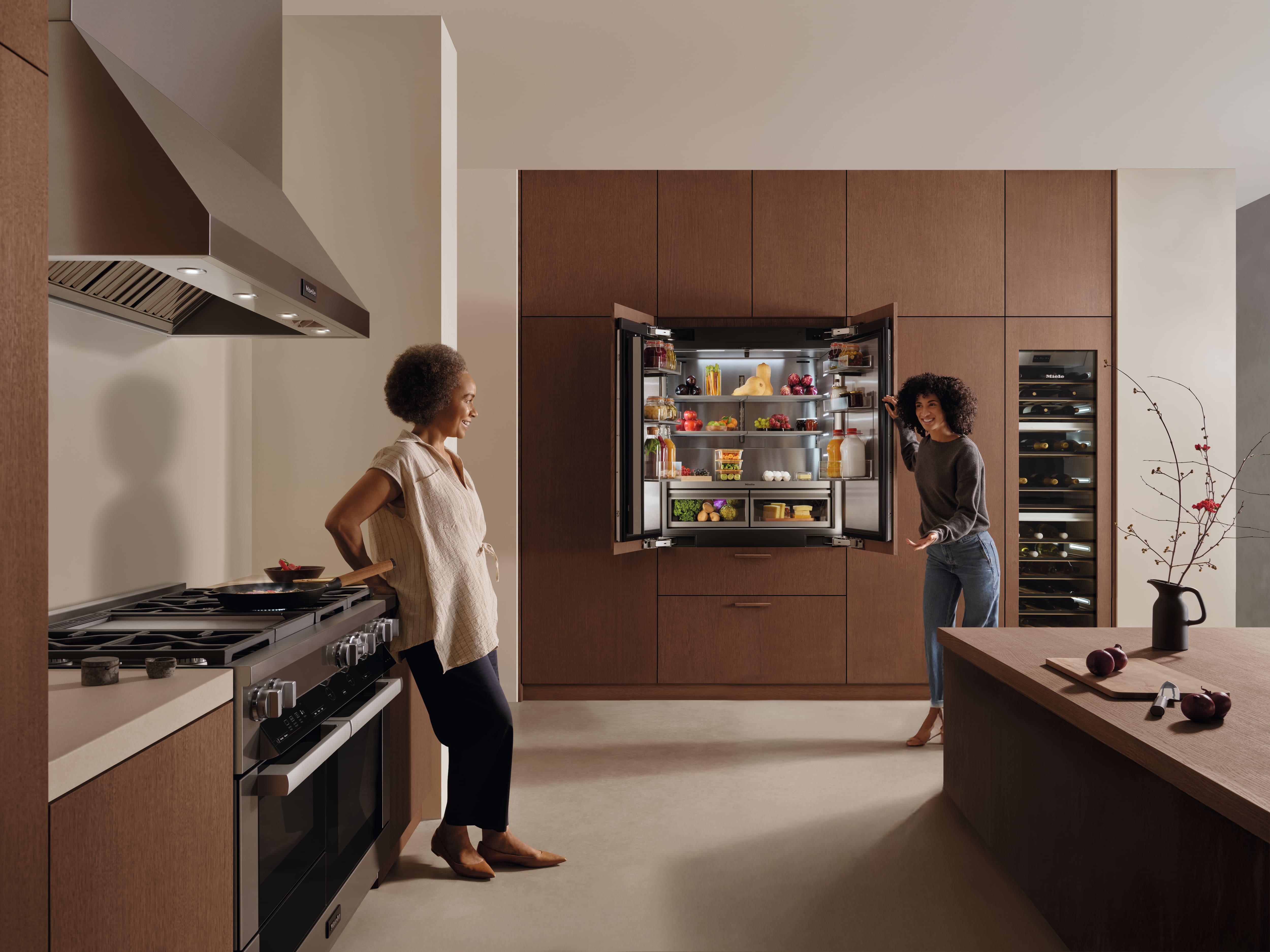 Cuisine moderne avec des meubles en bois ; une personne ouvre un réfrigérateur encastré rempli de provisions tandis qu'une autre est penchée près de la cuisinière.