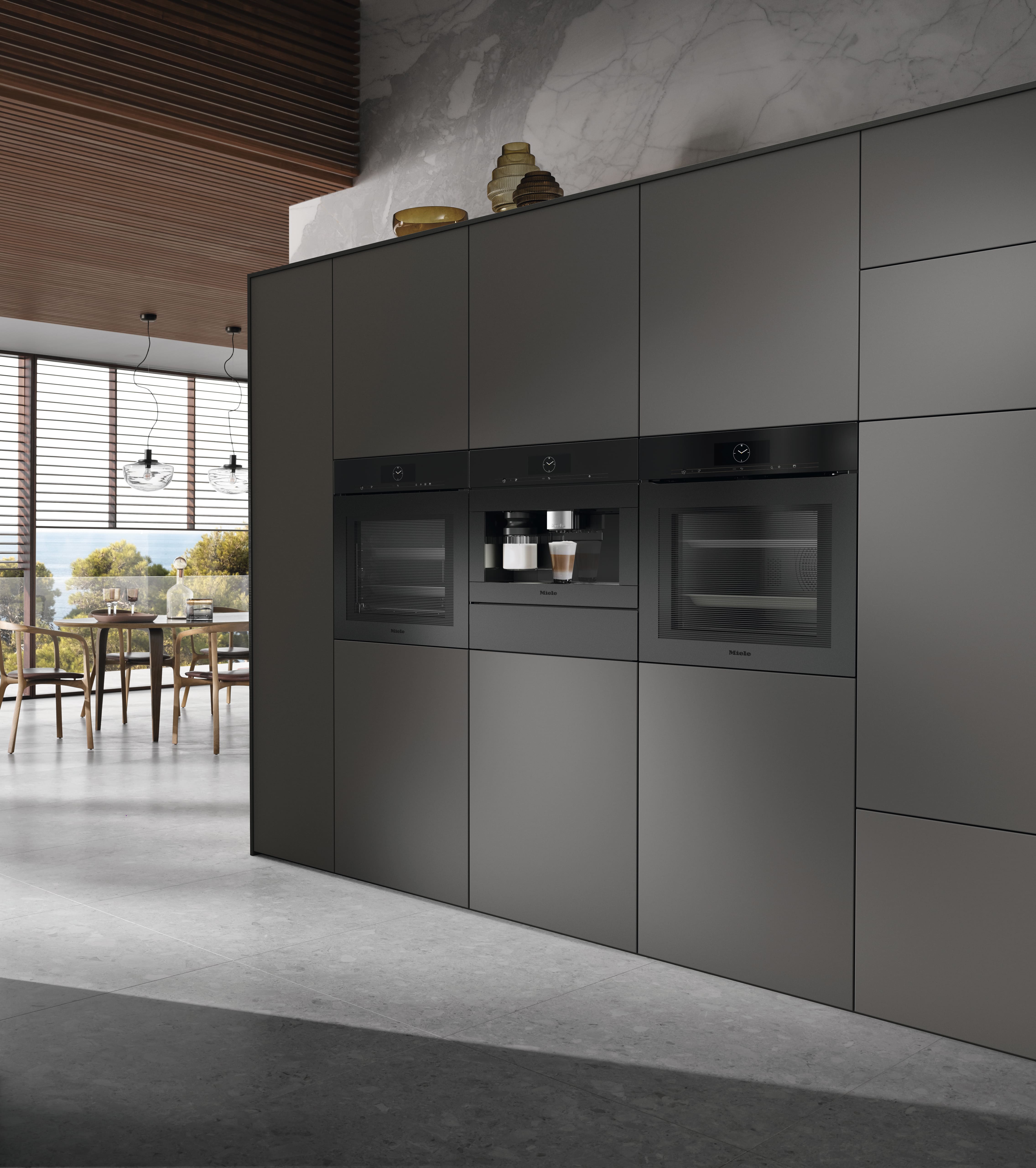 Sleek modern kitchen with Miele built‑in wall ovens and a Miele coffee machine, an open Miele dishwasher in a dark island, marble backsplash, floor‑to‑ceiling windows with blinds, and a dining area overlooking trees and water. Sleek modern kitchen with Miele built‑in wall ovens and a Miele coffee machine, an open Miele dishwasher in a dark island, marble backsplash, floor‑to‑ceiling windows with blinds, and a dining area overlooking trees and water.
