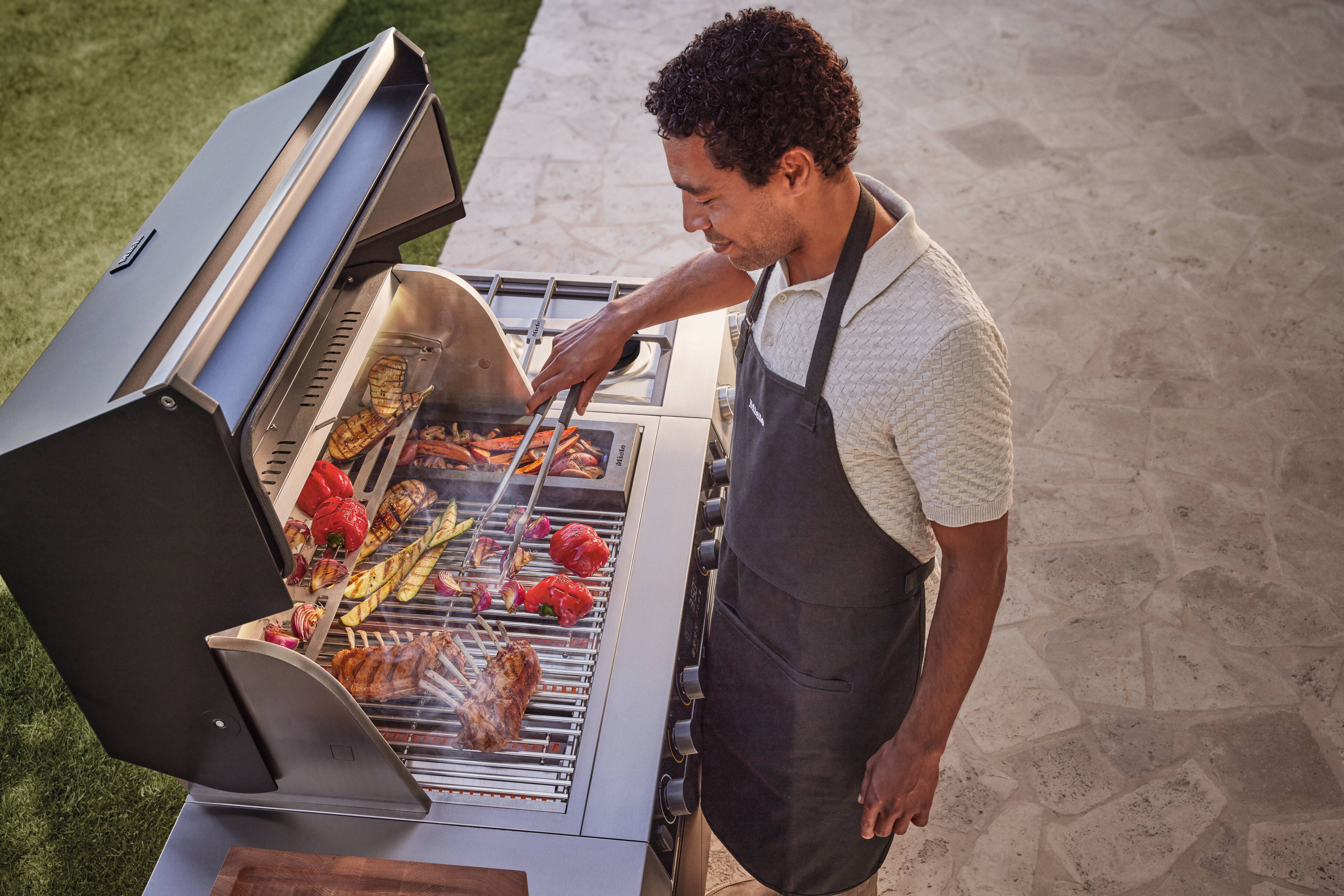 Person mit Schürze grillt verschiedenes Gemüse und Fleisch auf einem Edelstahl-Gasgrill auf einer Steinterrasse.