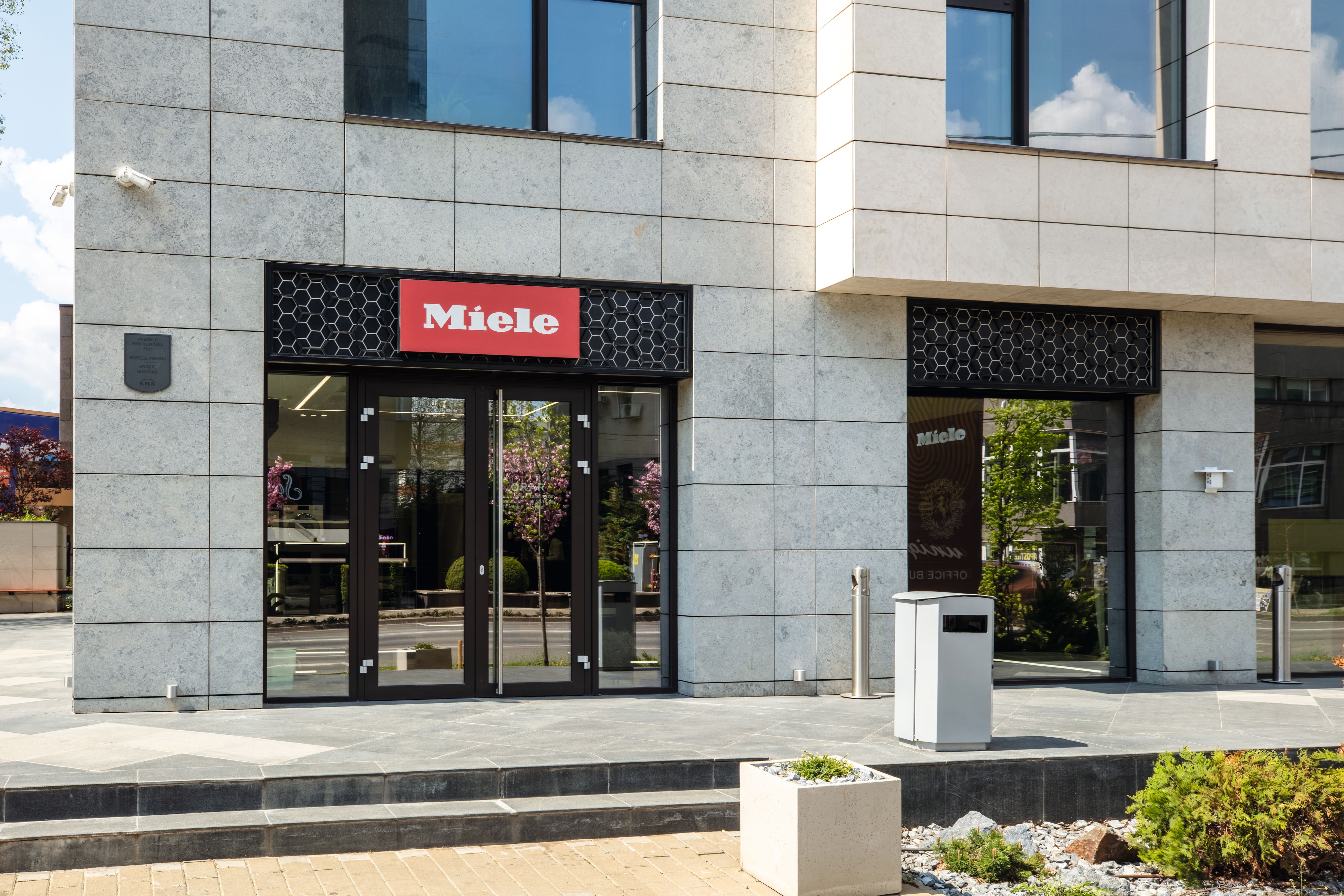 Clădire modernă cu fațadă din piatră, având la intrare un magazin Miele cu uși de sticlă, siglă roșie și trotuar amenajat. Clădire modernă cu fațadă din piatră, având la intrare un magazin Miele cu uși de sticlă, siglă roșie și trotuar amenajat.