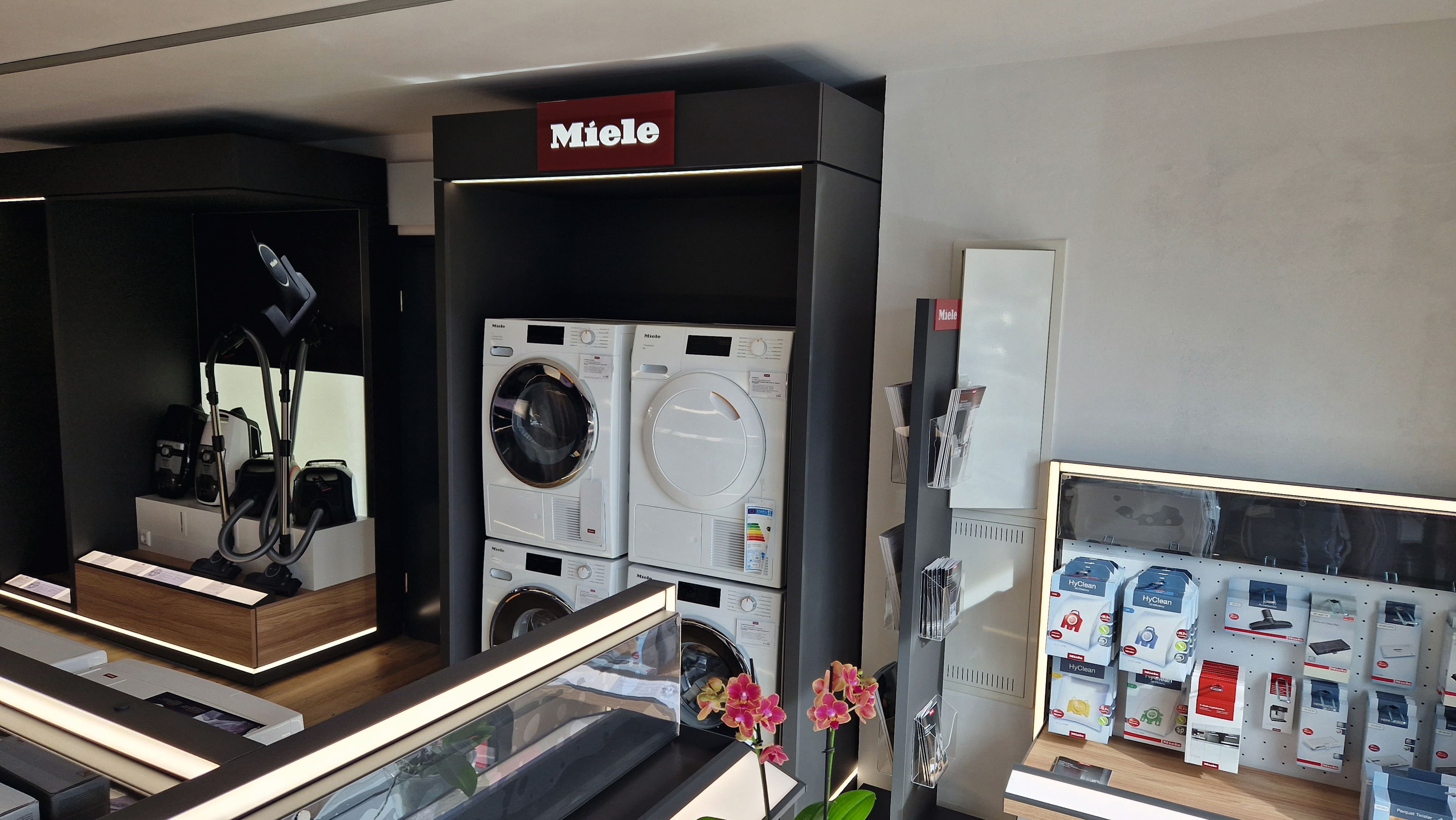 Ausstellungsfläche eines Elektrofachmarkts mit Miele-Logo, zwei gestapelte weiße Frontlader-Waschmaschinen und Trockner, Staubsauger links, Regale mit Miele HyClean-Zubehör rechts, moderne Beleuchtung und eine rosa Orchidee im Vordergrund.
