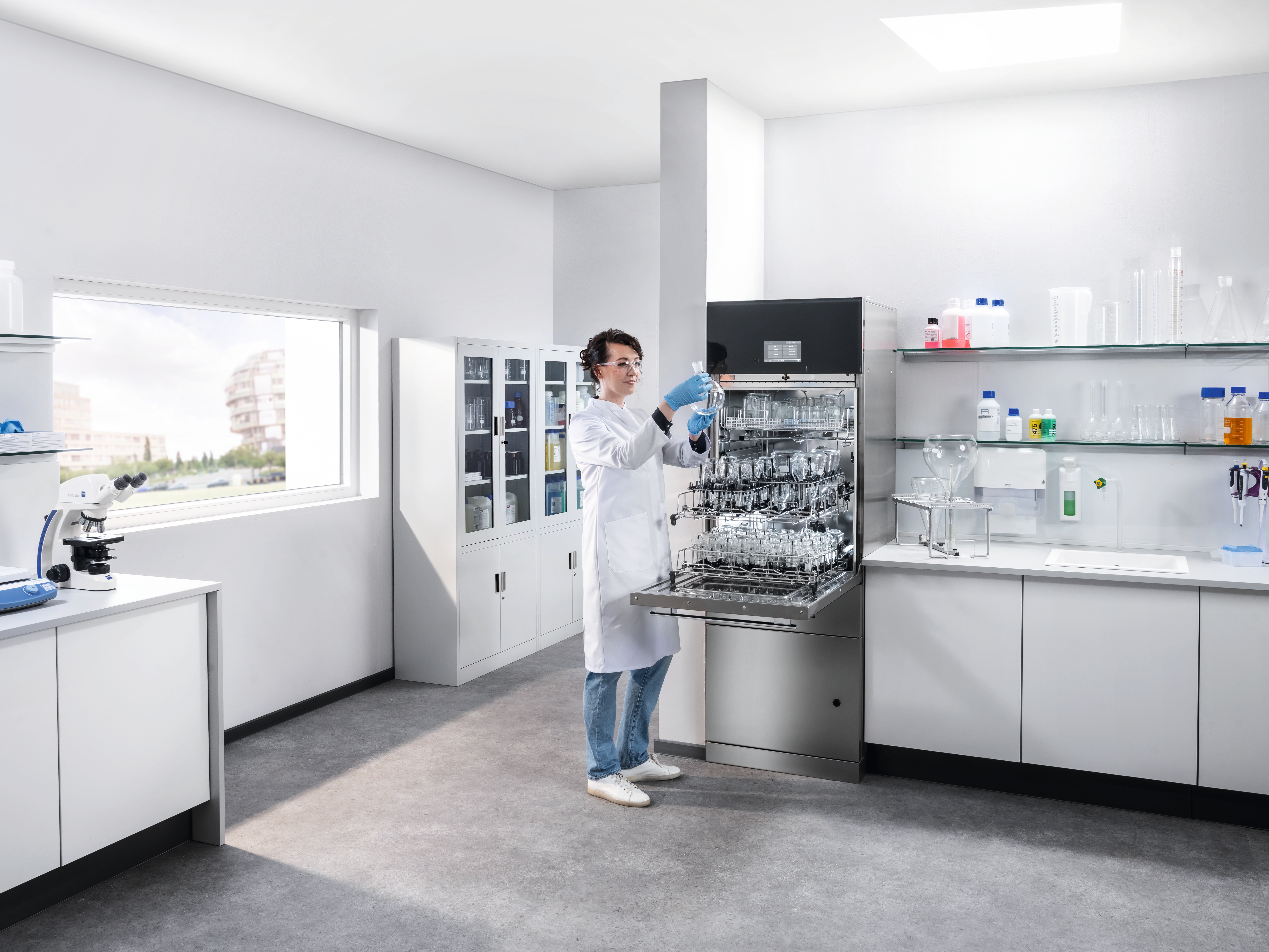 A lab technician is standing next to a SlimLine lab dishwasher, holding a clean round-bottom flask.