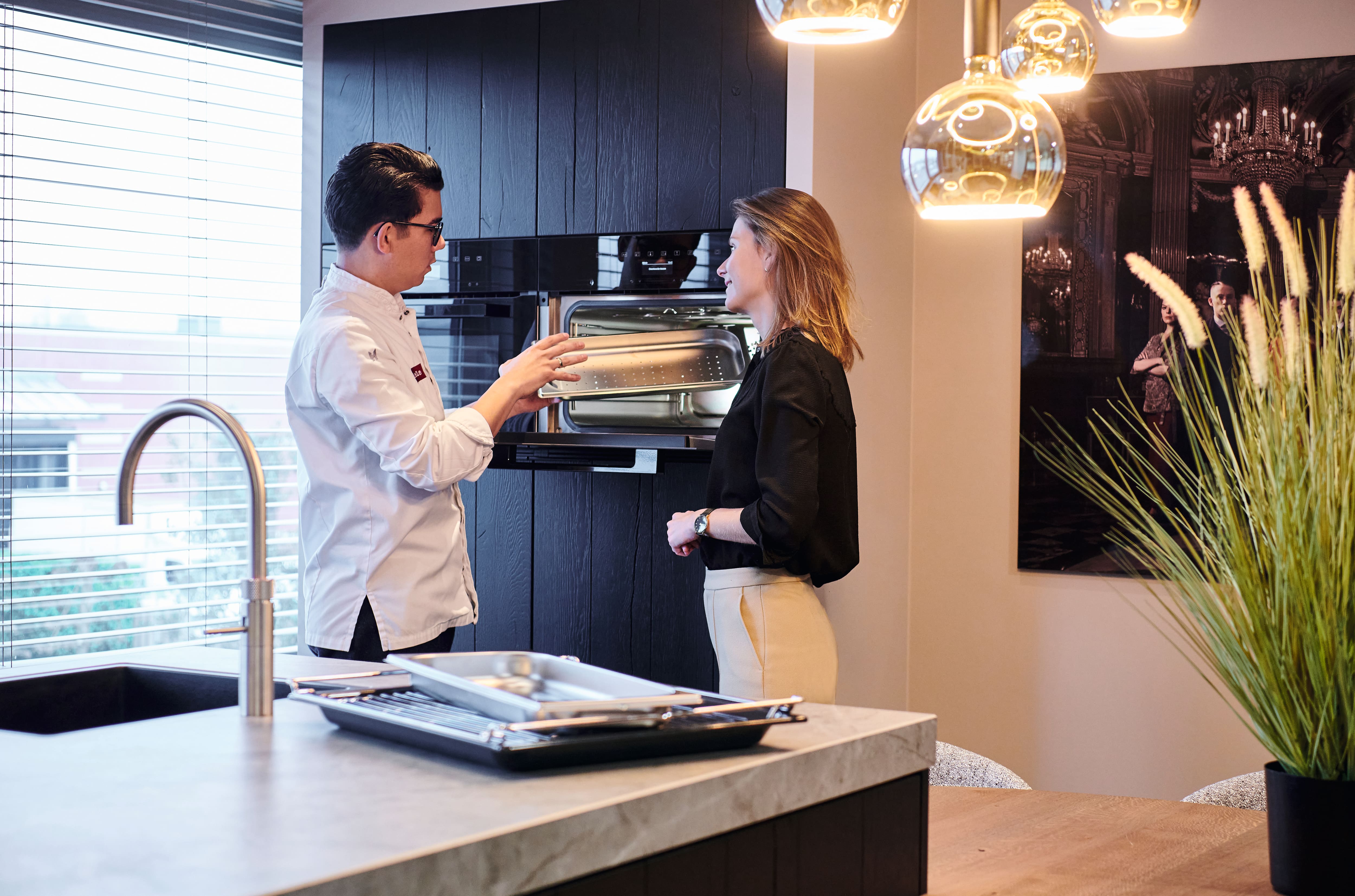 Modern kitchen showroom scene: staff member demonstrates a built-in steam oven and perforated tray to a visitor. Modern kitchen showroom scene: staff member demonstrates a built-in steam oven and perforated tray to a visitor.