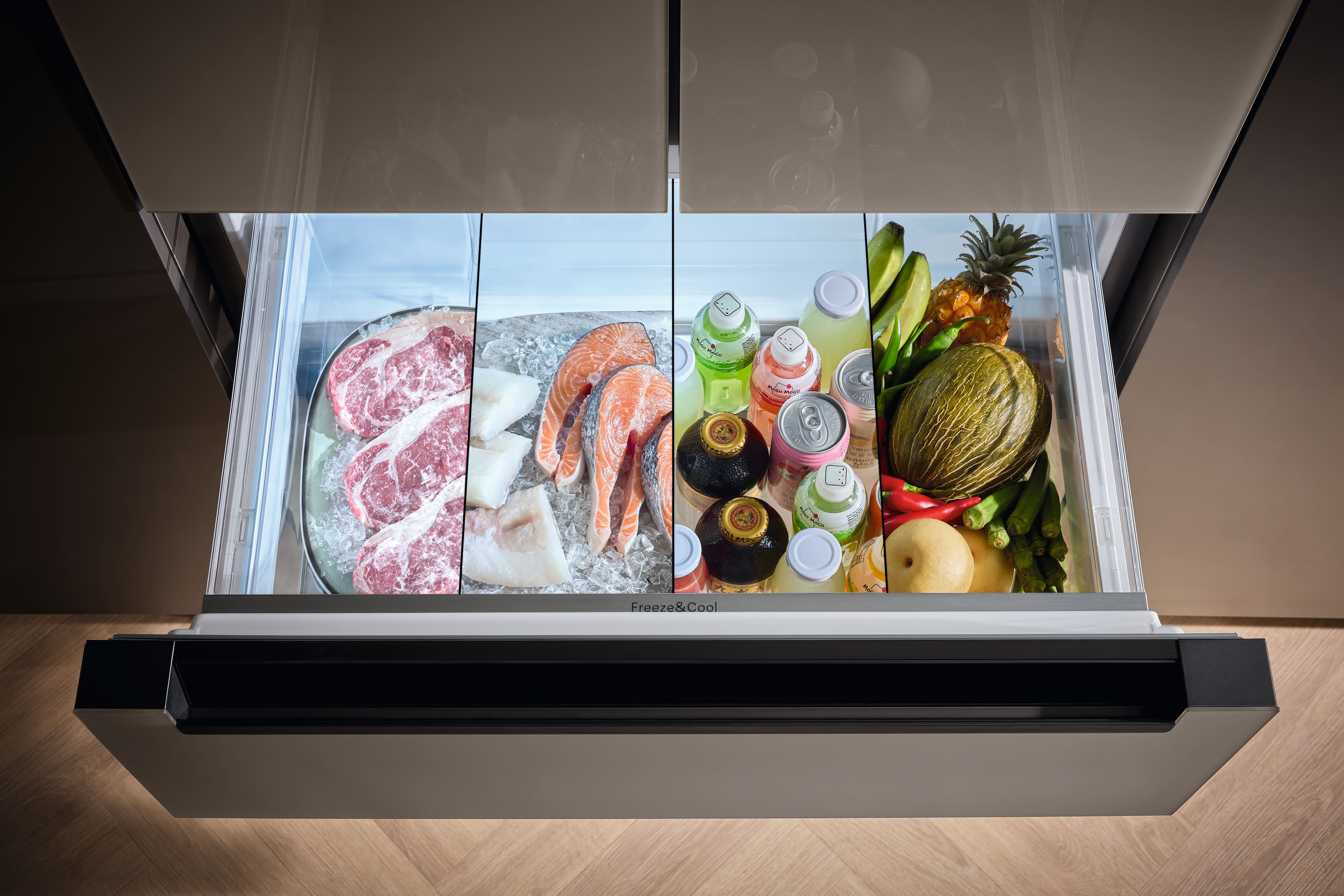 Open stainless steel refrigerator drawer showing a split compartment: left side with frosted plastic containers of frozen foods, right side with cans and bottles of drinks and a decorated cake. Open stainless steel refrigerator drawer showing a split compartment: left side with frosted plastic containers of frozen foods, right side with cans and bottles of drinks and a decorated cake.