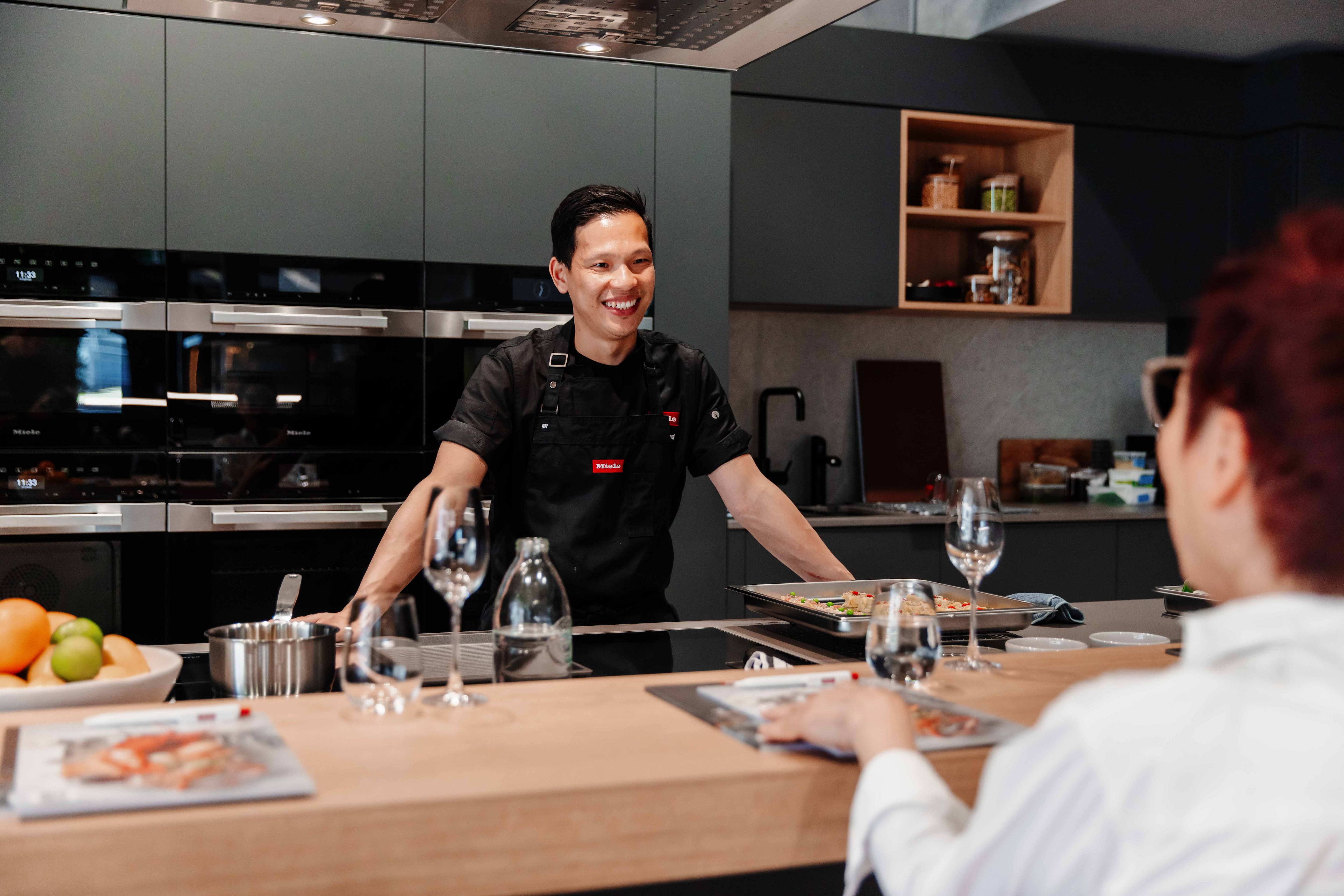 Smiling chef leads a cooking demo in a modern kitchen, standing at an island with ovens, glassware, and a tray of food.
