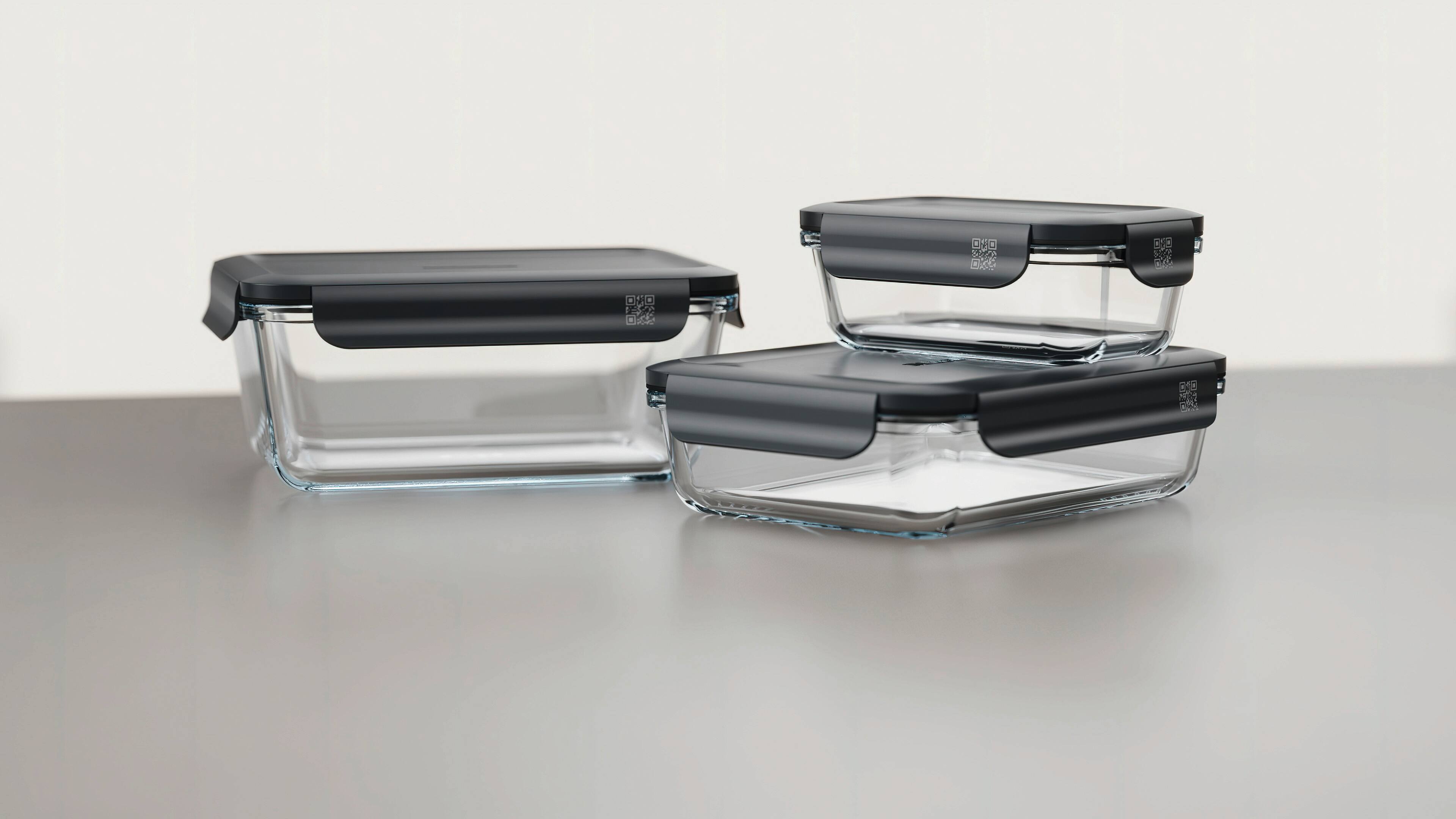 Set of clear glass food storage containers with dark gray locking lids, shown stacked on a counter for kitchen meal prep. Set of clear glass food storage containers with dark gray locking lids, shown stacked on a counter for kitchen meal prep.