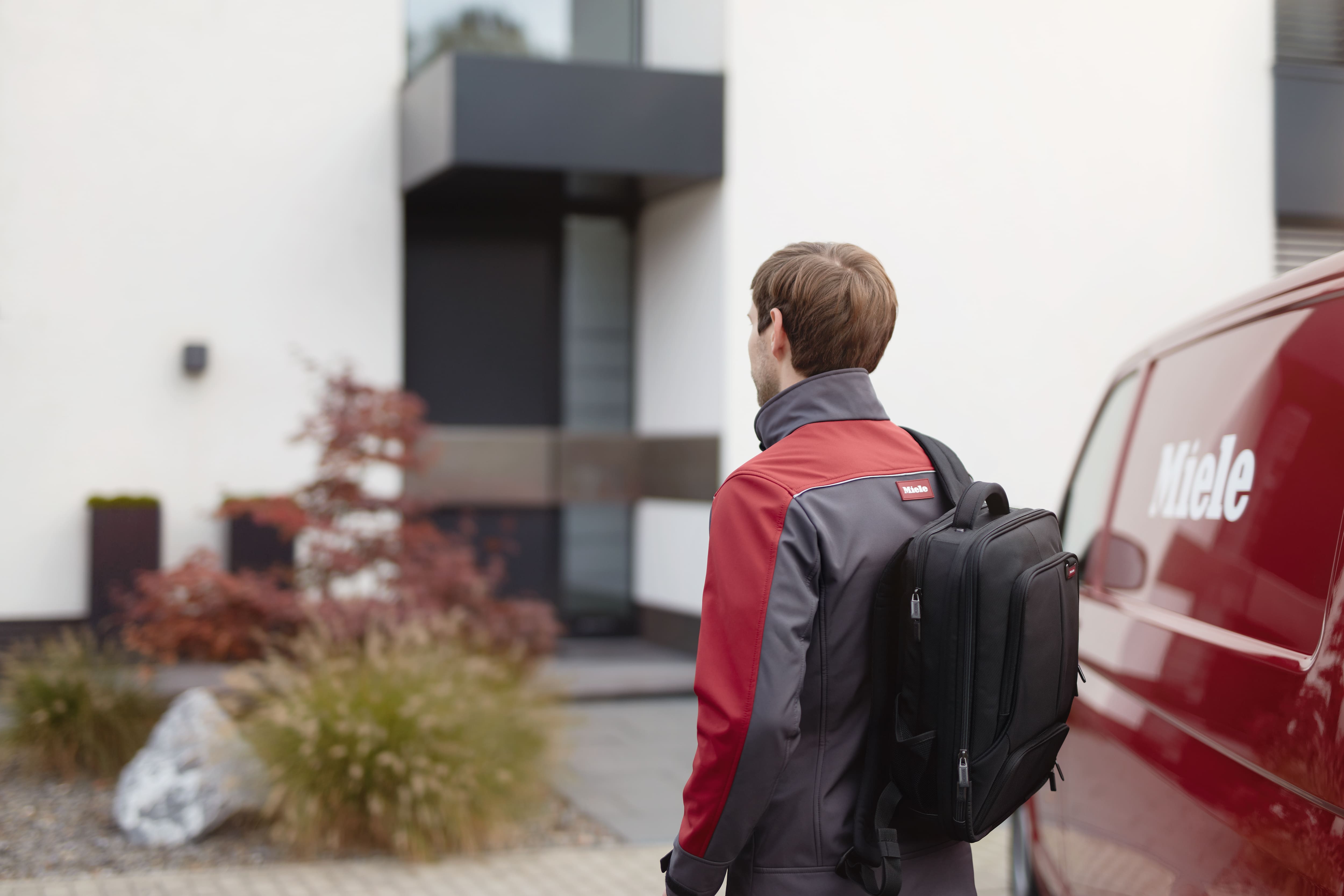 Miele Servicetechniker auf dem Weg zu einem modernen Wohnhaus neben einem Miele Servicefahrzeug. Das Bild symbolisiert Kundendienst und technischen Support vor Ort.