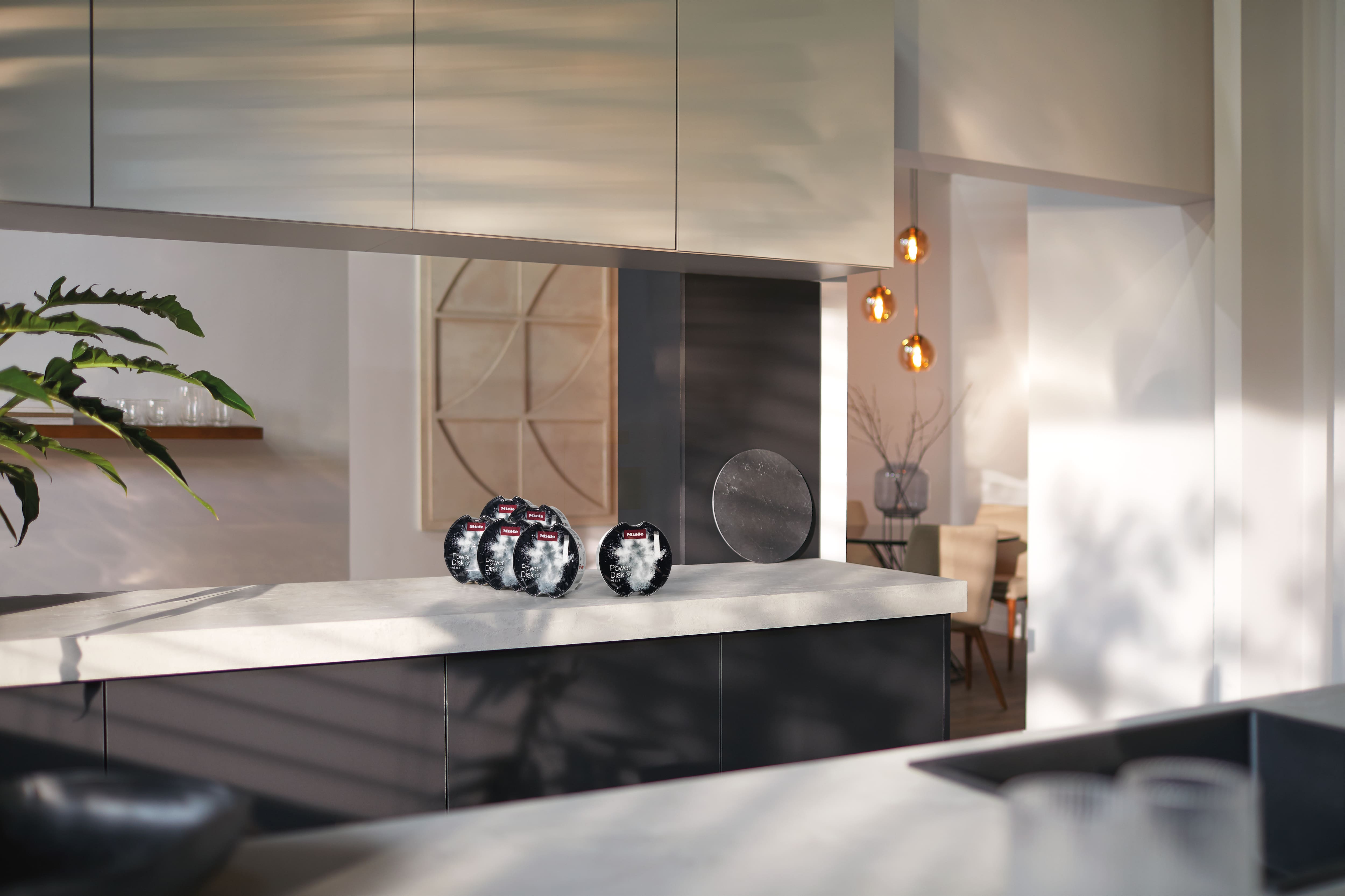 Contemporary kitchen with light countertop; cluster of round black product packs on island, warm pendant lights in dining area.