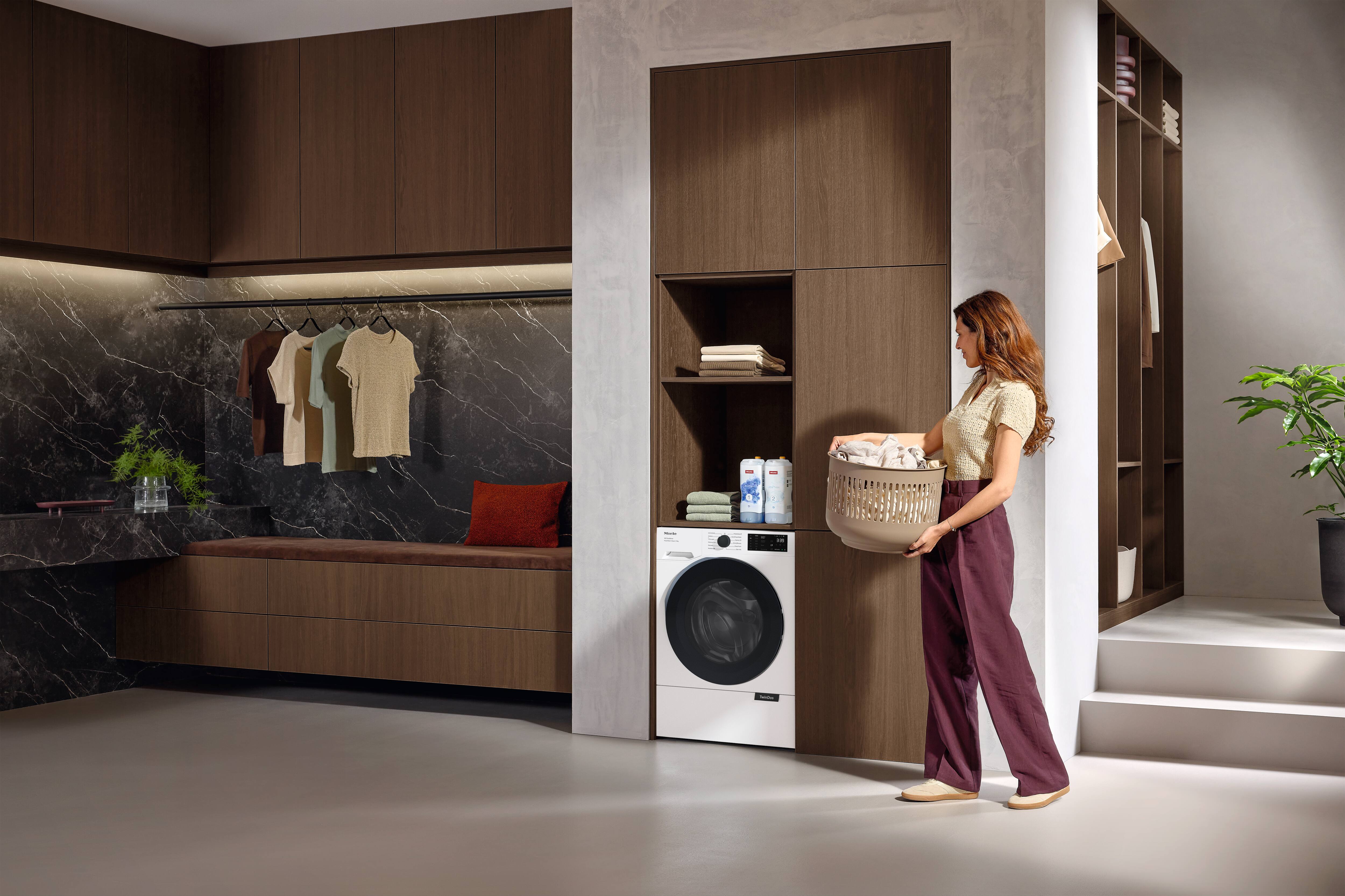 Modern laundry room with built-in front-load washer, warm wood cabinetry, and a woman carrying a laundry basket. Modern laundry room with built-in front-load washer, warm wood cabinetry, and a woman carrying a laundry basket.
