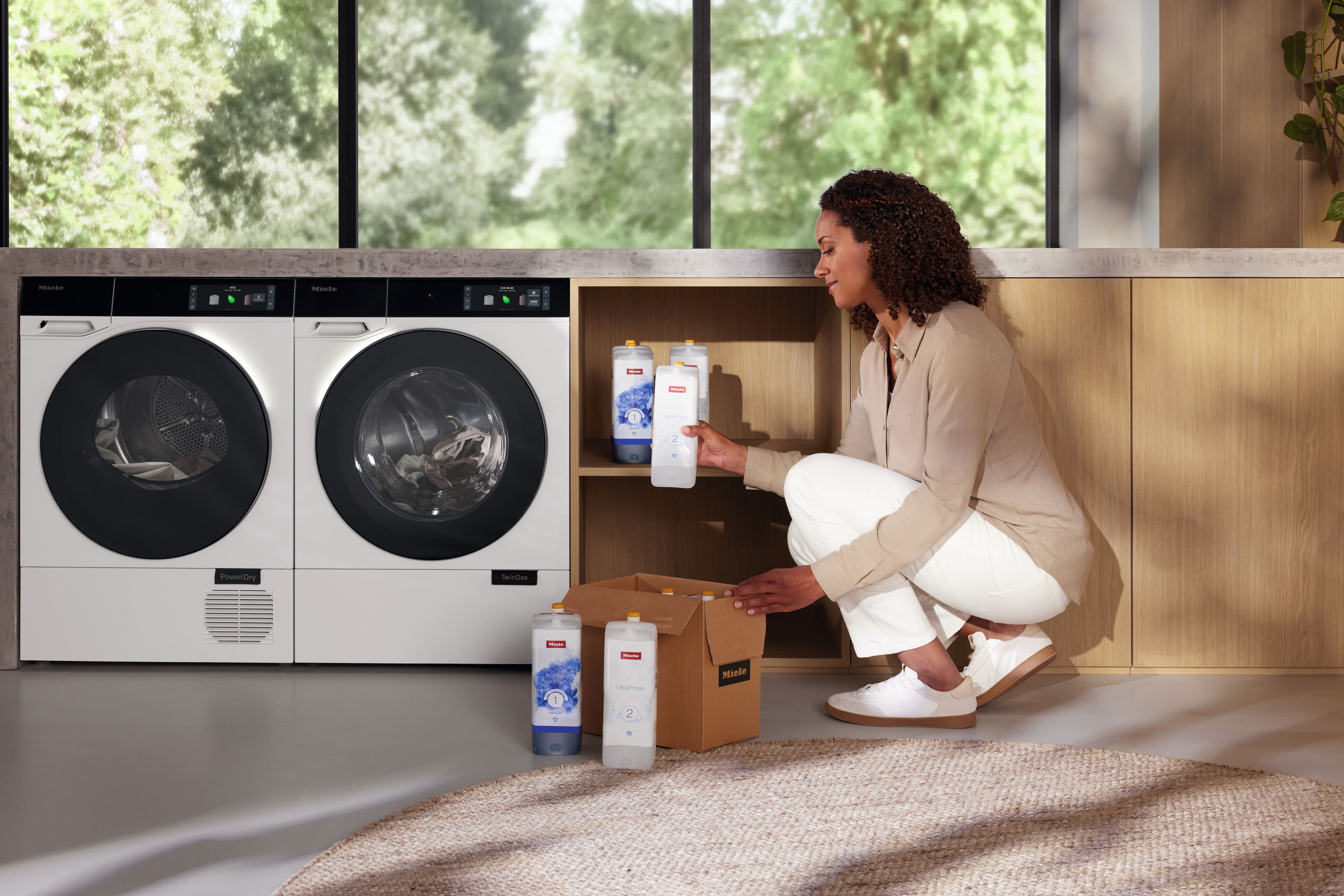 Une personne range les cartouches de lessive Miele TwinDos à côté d'un lave-linge et d'un sèche-linge assortis dans une buanderie moderne Une personne range les cartouches de lessive Miele TwinDos à côté d'un lave-linge et d'un sèche-linge assortis dans une buanderie moderne