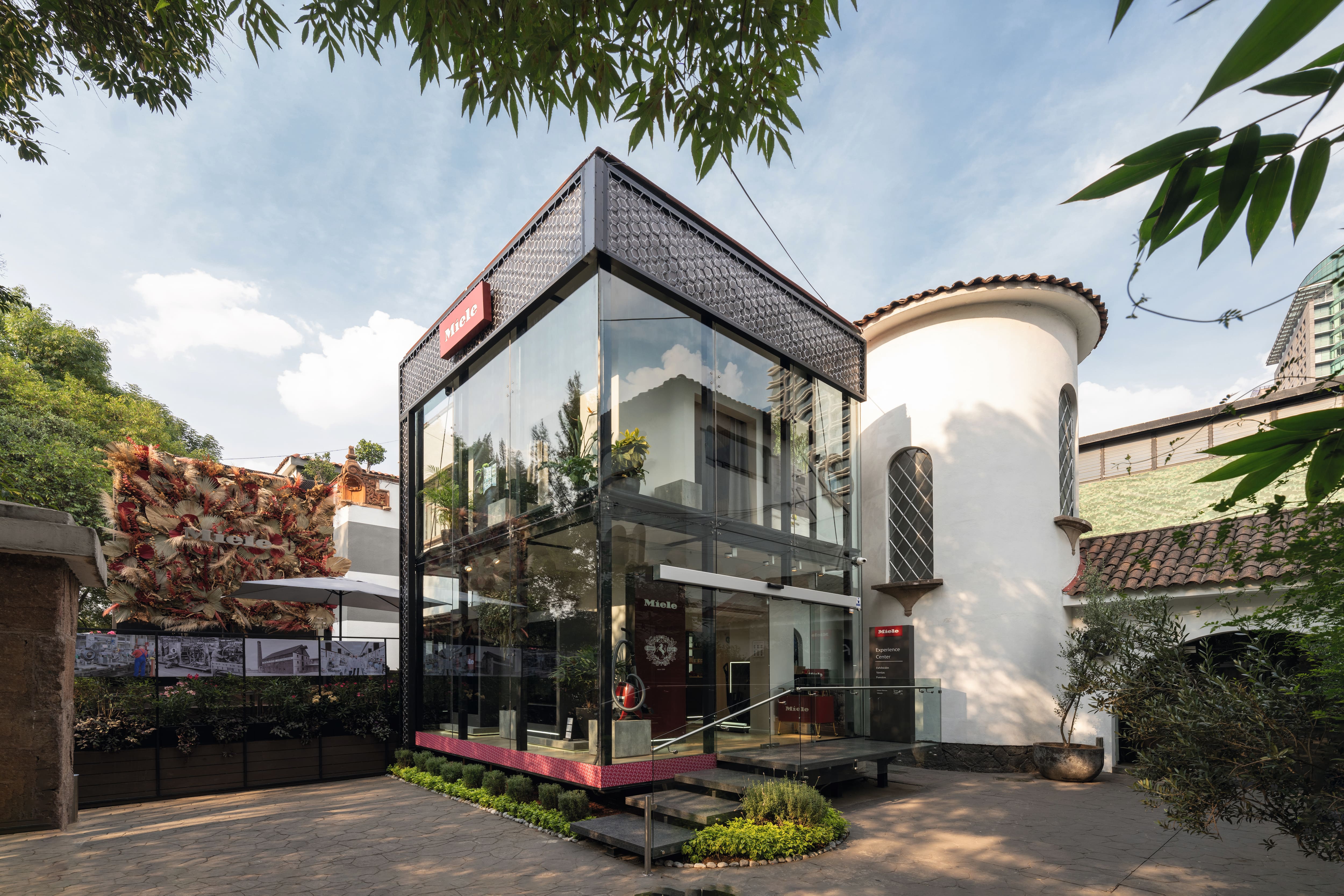 Sala de exhibición moderna de vidrio con la marca Miele junto a una torre de estuco blanco, ubicada en un patio arbolado.