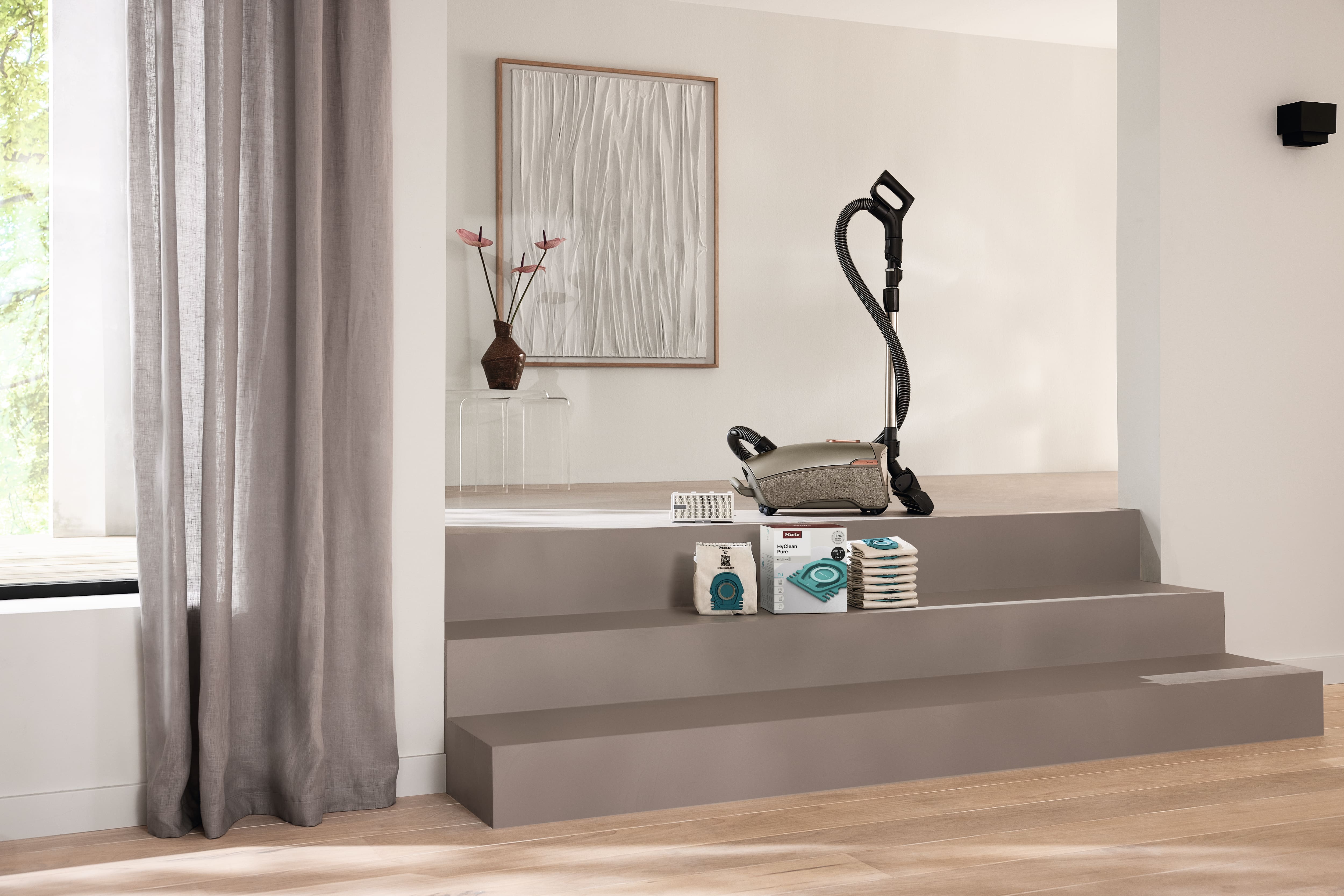 Interno moderno con un aspirapolvere a traino su scale color tortora, circondato da sacchetti per aspirapolvere e filtri, vicino a tende trasparenti e a un’opera d’arte astratta incorniciata. Interno moderno con un aspirapolvere a traino su scale color tortora, circondato da sacchetti per aspirapolvere e filtri, vicino a tende trasparenti e a un’opera d’arte astratta incorniciata.