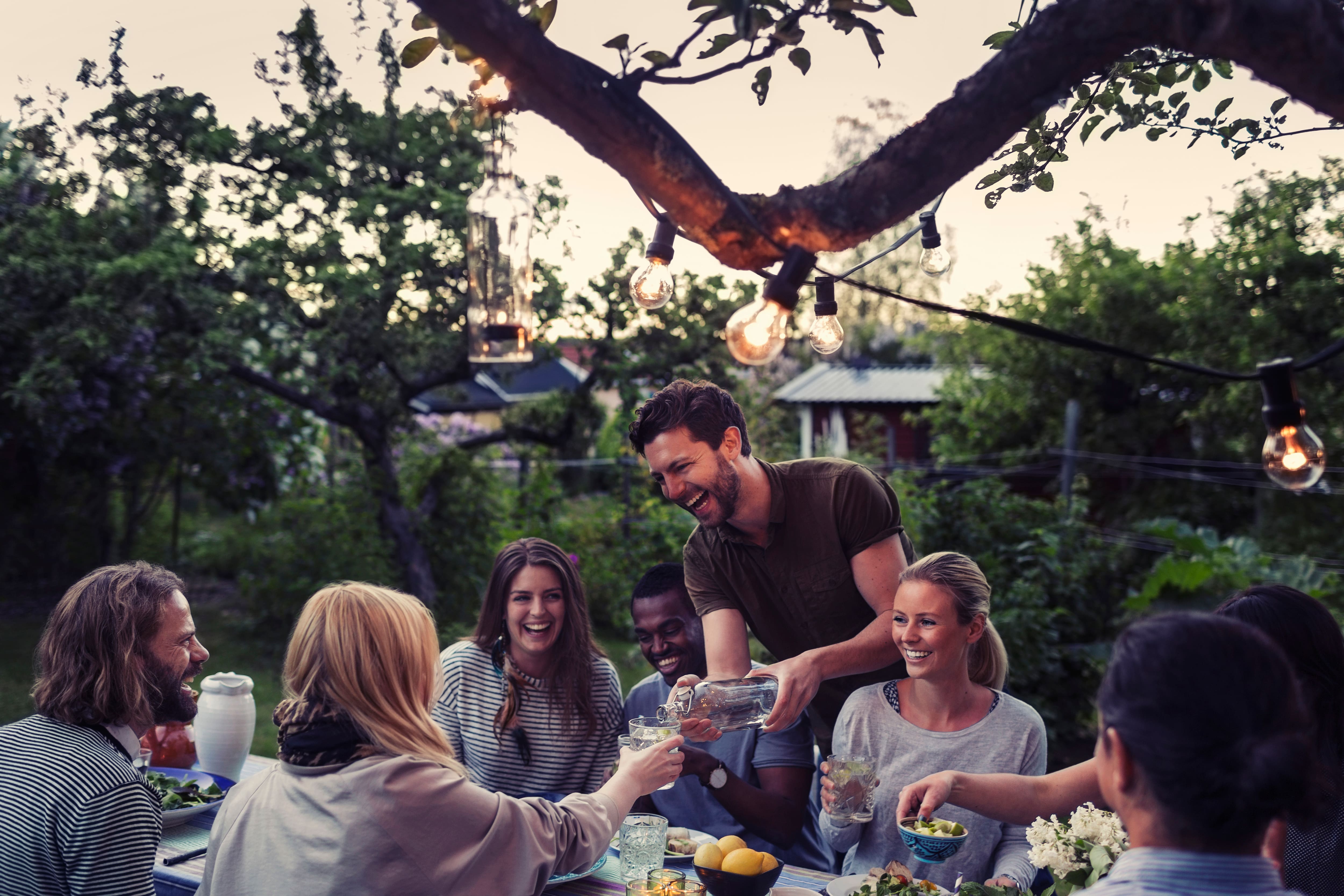 Degli amici gustano una cena all’aperto al crepuscolo, sotto le lucine, mentre un uomo versa dell’acqua al tavolo da giardino. Degli amici gustano una cena all’aperto al crepuscolo, sotto le lucine, mentre un uomo versa dell’acqua al tavolo da giardino.