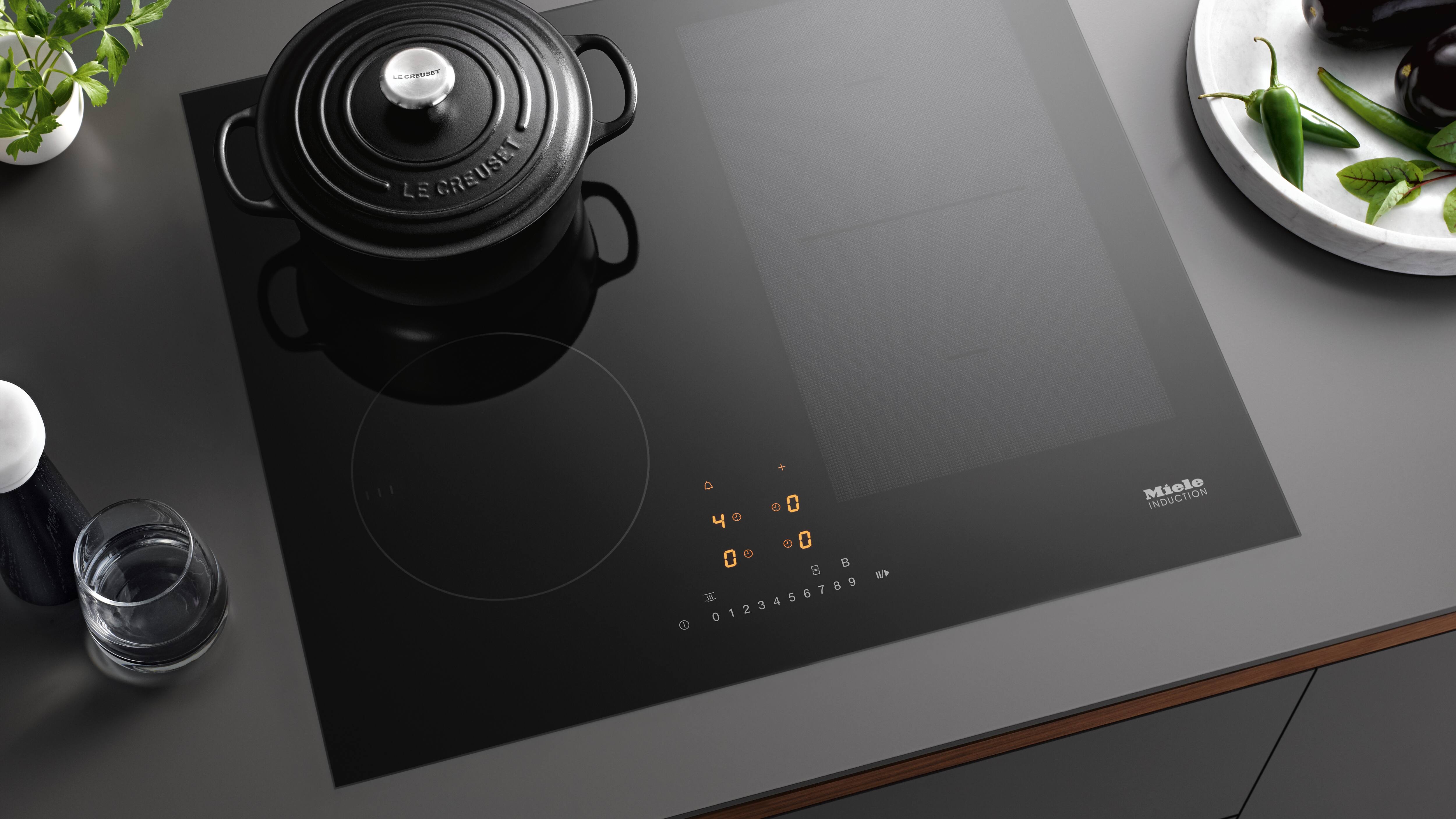 Modern black induction cooktop with illuminated touch controls, a black Le Creuset Dutch oven on the left burner, and nearby plate of vegetables; Miele Induction branding visible. Modern black induction cooktop with illuminated touch controls, a black Le Creuset Dutch oven on the left burner, and nearby plate of vegetables; Miele Induction branding visible.