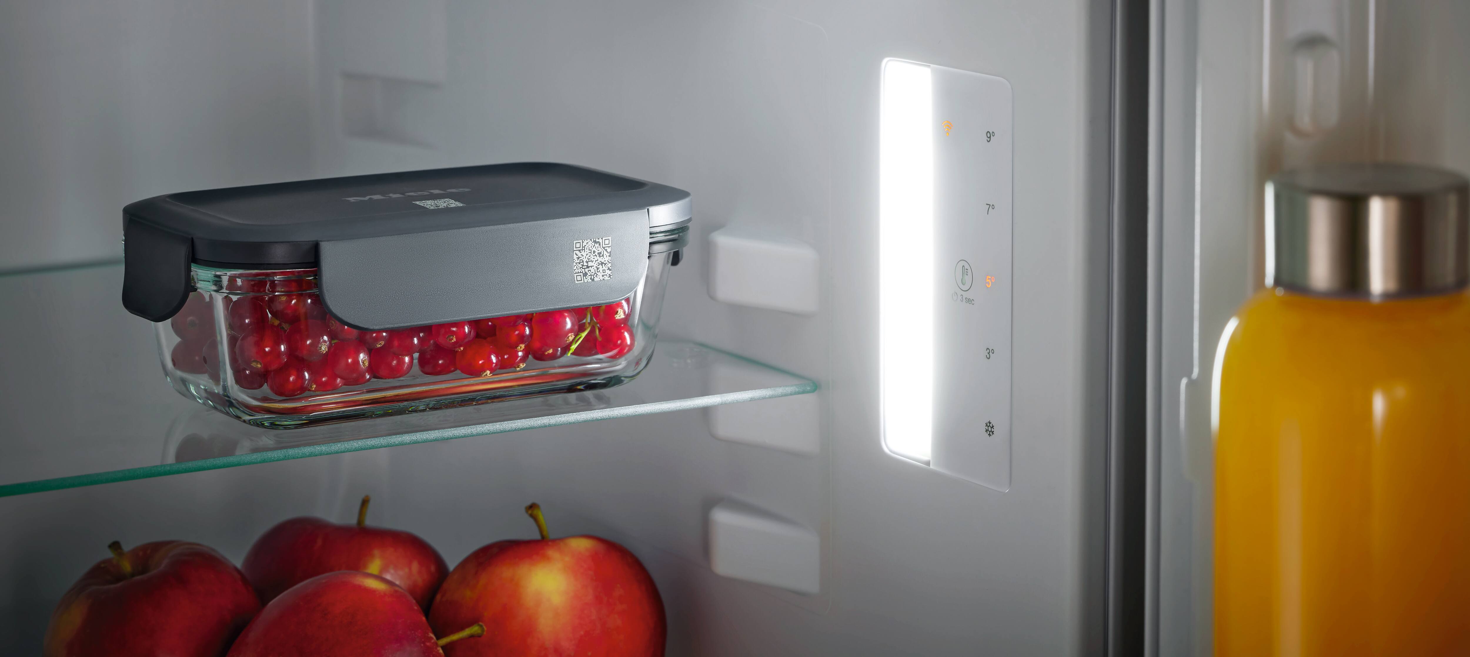 Inside a refrigerator, a glass storage container with a dark gray lid and a QR code label holds red berries on a glass shelf; apples sit on the lower shelf, an illuminated control panel shows temperature settings, and an orange drink bottle is in the door. Inside a refrigerator, a glass storage container with a dark gray lid and a QR code label holds red berries on a glass shelf; apples sit on the lower shelf, an illuminated control panel shows temperature settings, and an orange drink bottle is in the door.