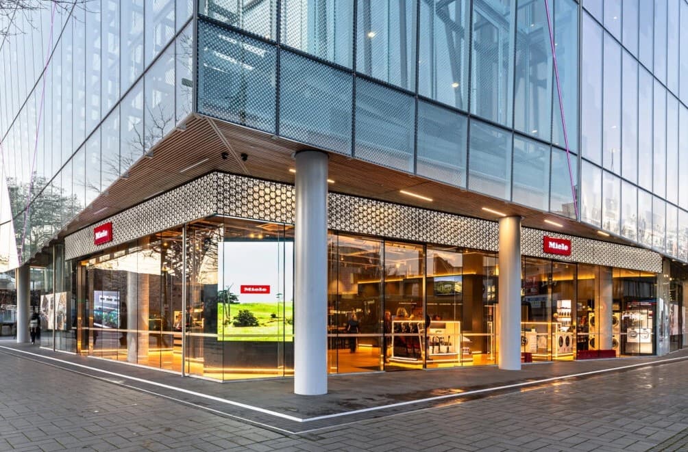 Modern glass-walled Miele appliance showroom at a street corner, warm interior lighting and product displays visible.