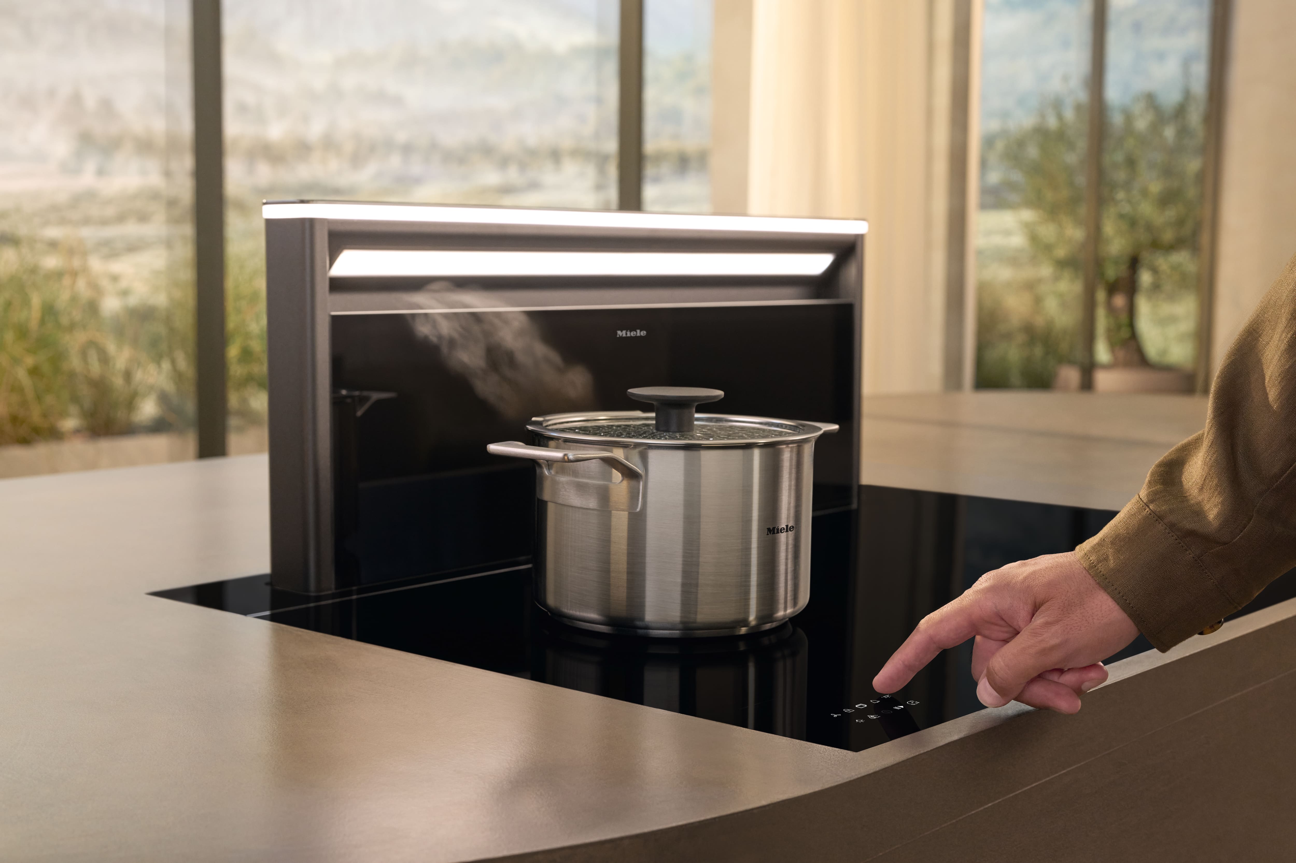Modern kitchen with Miele induction cooktop and downdraft hood; hand adjusts touch controls as a pot steams. Modern kitchen with Miele induction cooktop and downdraft hood; hand adjusts touch controls as a pot steams.