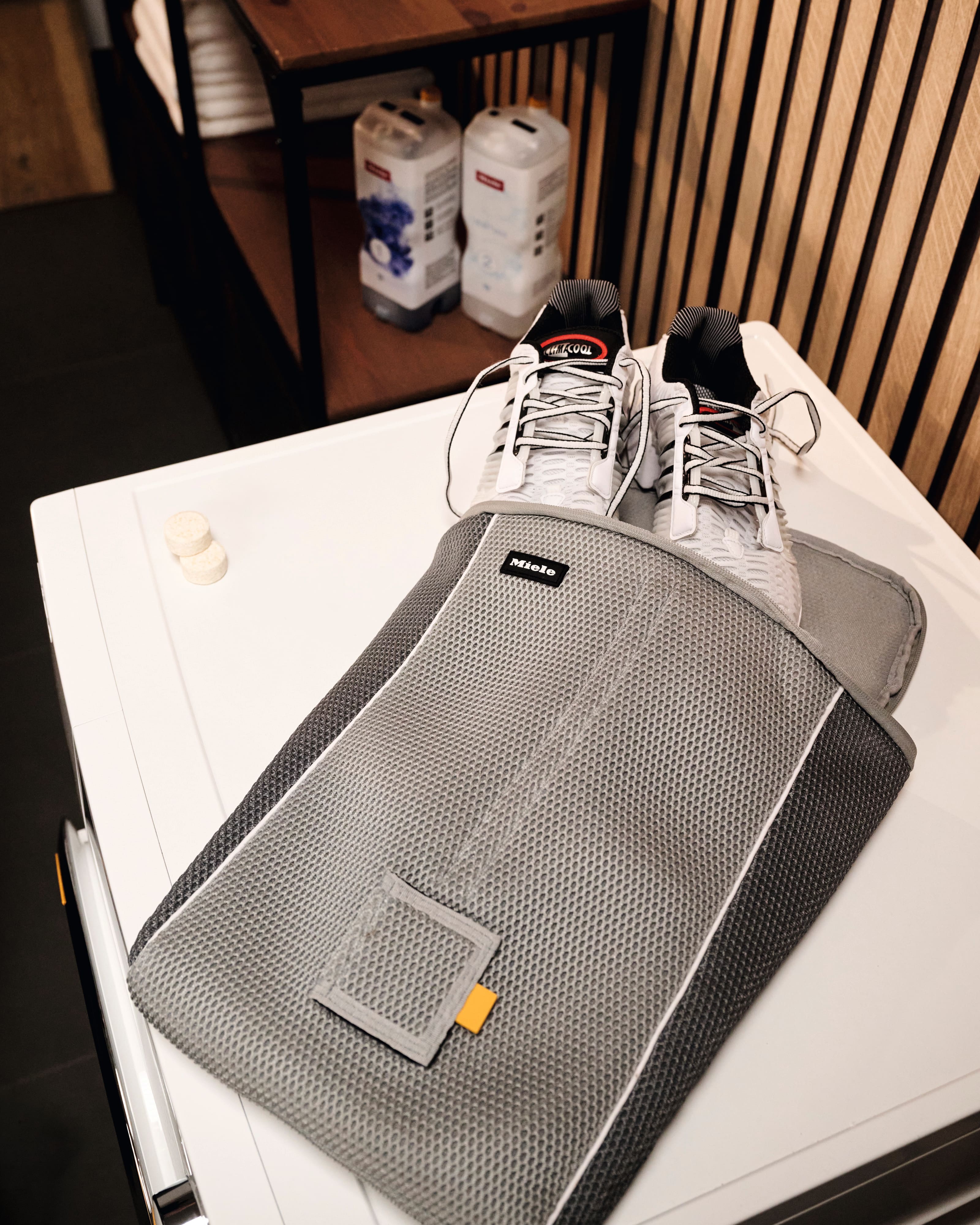 Offener grauer Schuhorganizer aus Mesh mit gepolsterten Fächern auf einem Holzregal; daneben weisse Sneaker.