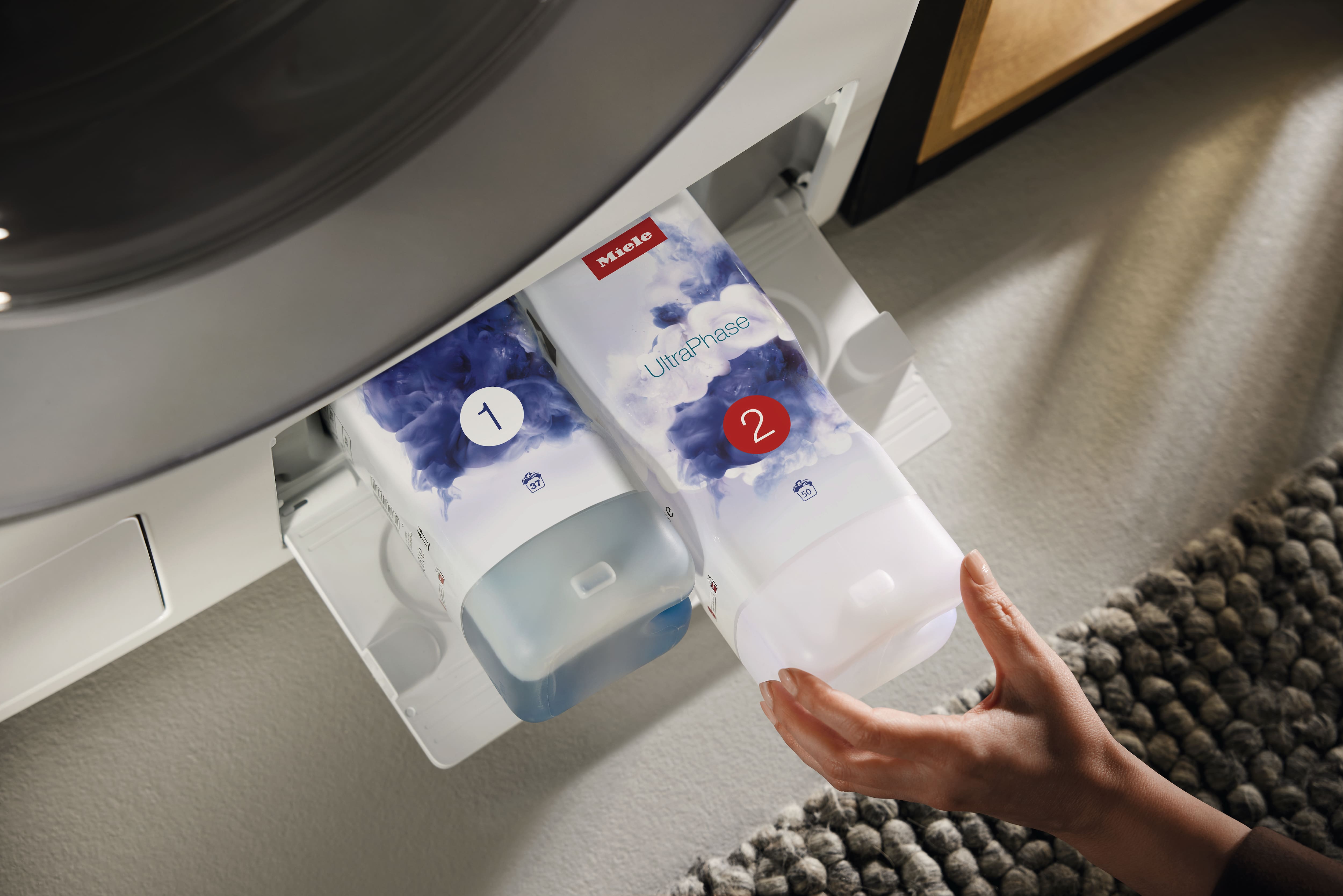 Close-up of a washing machine’s TwinDos drawer with two Miele UltraPhase detergent cartridges labeled 1 and 2, a hand sliding the “2” bottle into place. Close-up of a washing machine’s TwinDos drawer with two Miele UltraPhase detergent cartridges labeled 1 and 2, a hand sliding the “2” bottle into place.