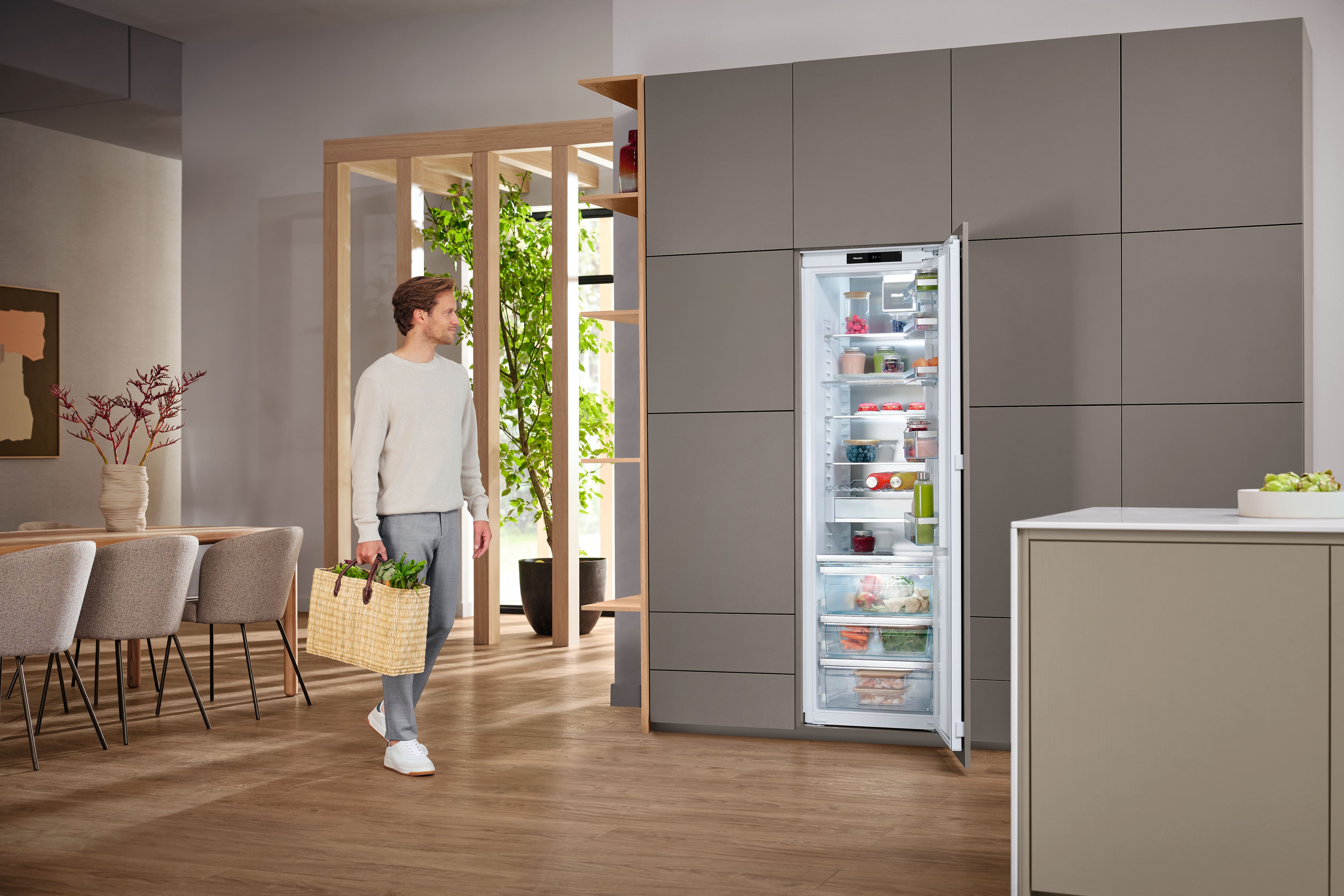 Eine Person trägt einen geflochtenen Einkaufskorb mit frischem Obst und Gemüse in eine moderne Küche und geht auf einen geöffneten Einbau-Kühlschrank zu. Graue Schränke, Holzboden, ein Esstisch und Zimmerpflanzen ergänzen die Szene. Eine Person trägt einen geflochtenen Einkaufskorb mit frischem Obst und Gemüse in eine moderne Küche und geht auf einen geöffneten Einbau-Kühlschrank zu. Graue Schränke, Holzboden, ein Esstisch und Zimmerpflanzen ergänzen die Szene.
