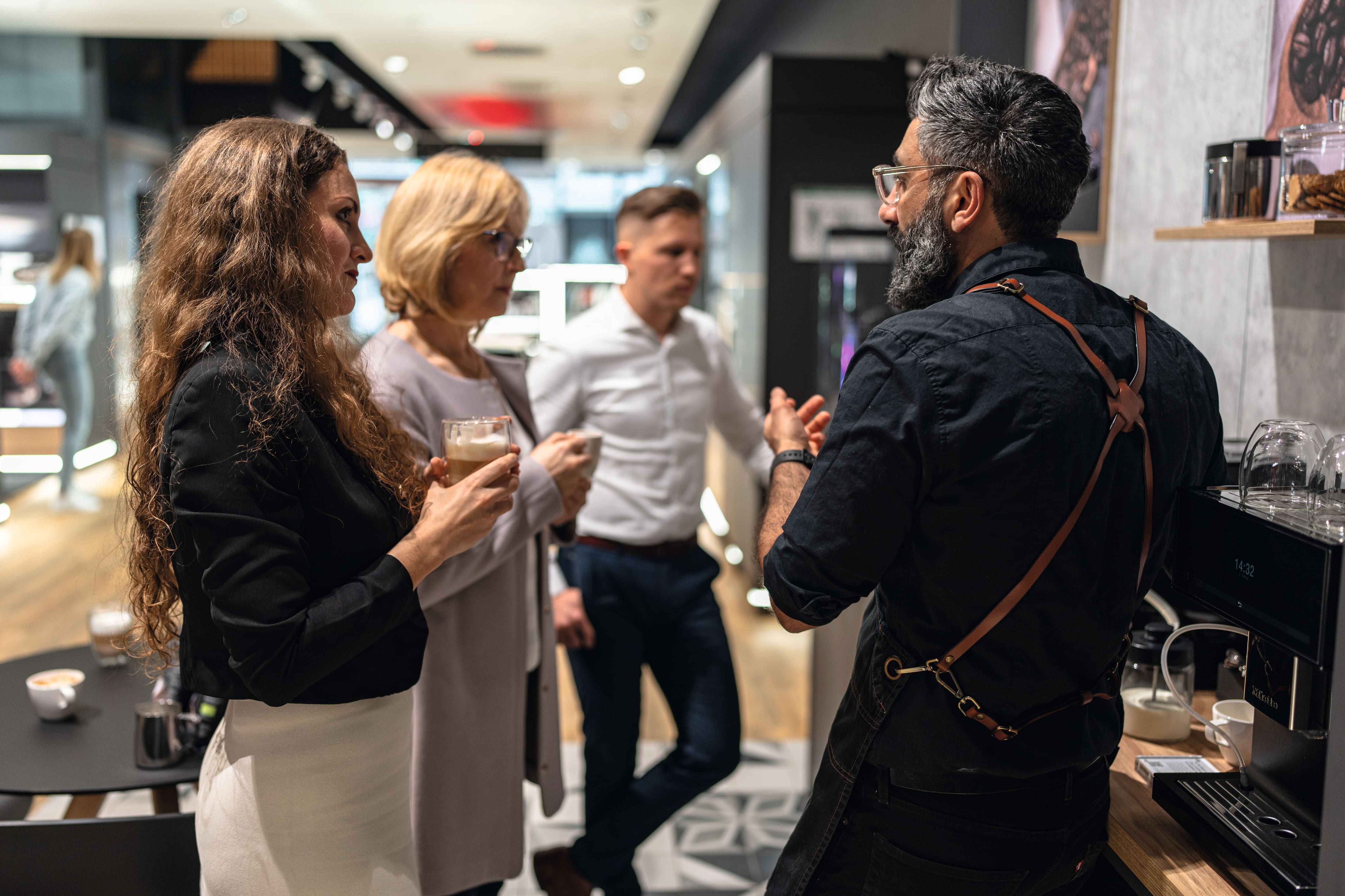 Barista purtând un șorț cu curea de piele explică prepararea cafelei la trei adulți cu latte-uri într-un showroom modern de cafenea lângă un espressor. Barista purtând un șorț cu curea de piele explică prepararea cafelei la trei adulți cu latte-uri într-un showroom modern de cafenea lângă un espressor.