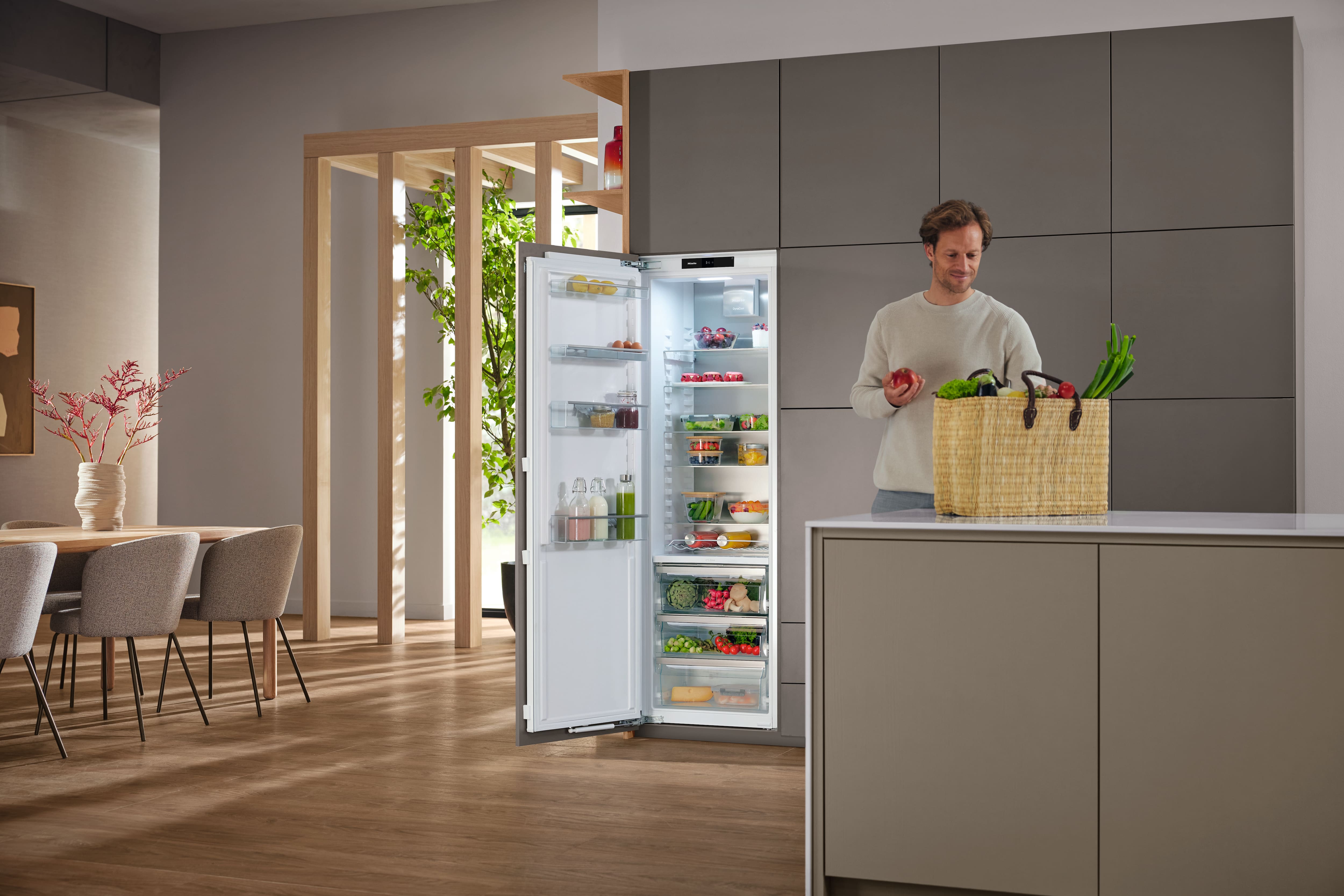 Modern kitchen interior with an open built‑in refrigerator full of food; a person stands by the island unpacking a wicker basket of fresh produce; gray cabinetry, wooden floors, and plants visible in natural light.