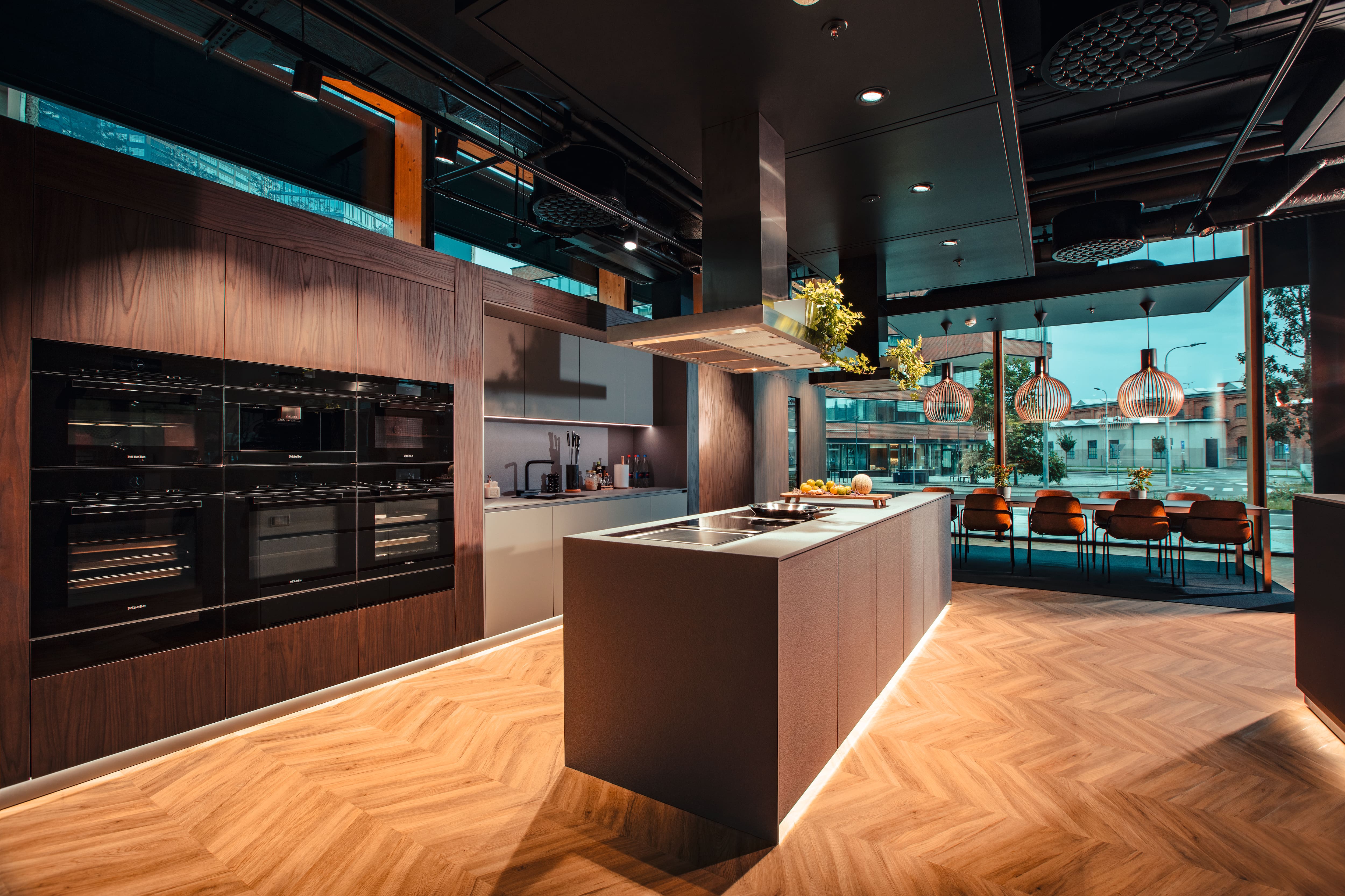 Moderní showroom kuchyně s ostrůvkem, vestavěnými troubami, dřevěnou podlahou a jídelnou se skleněnými stěnami. Moderní showroom kuchyně s ostrůvkem, vestavěnými troubami, dřevěnou podlahou a jídelnou se skleněnými stěnami.