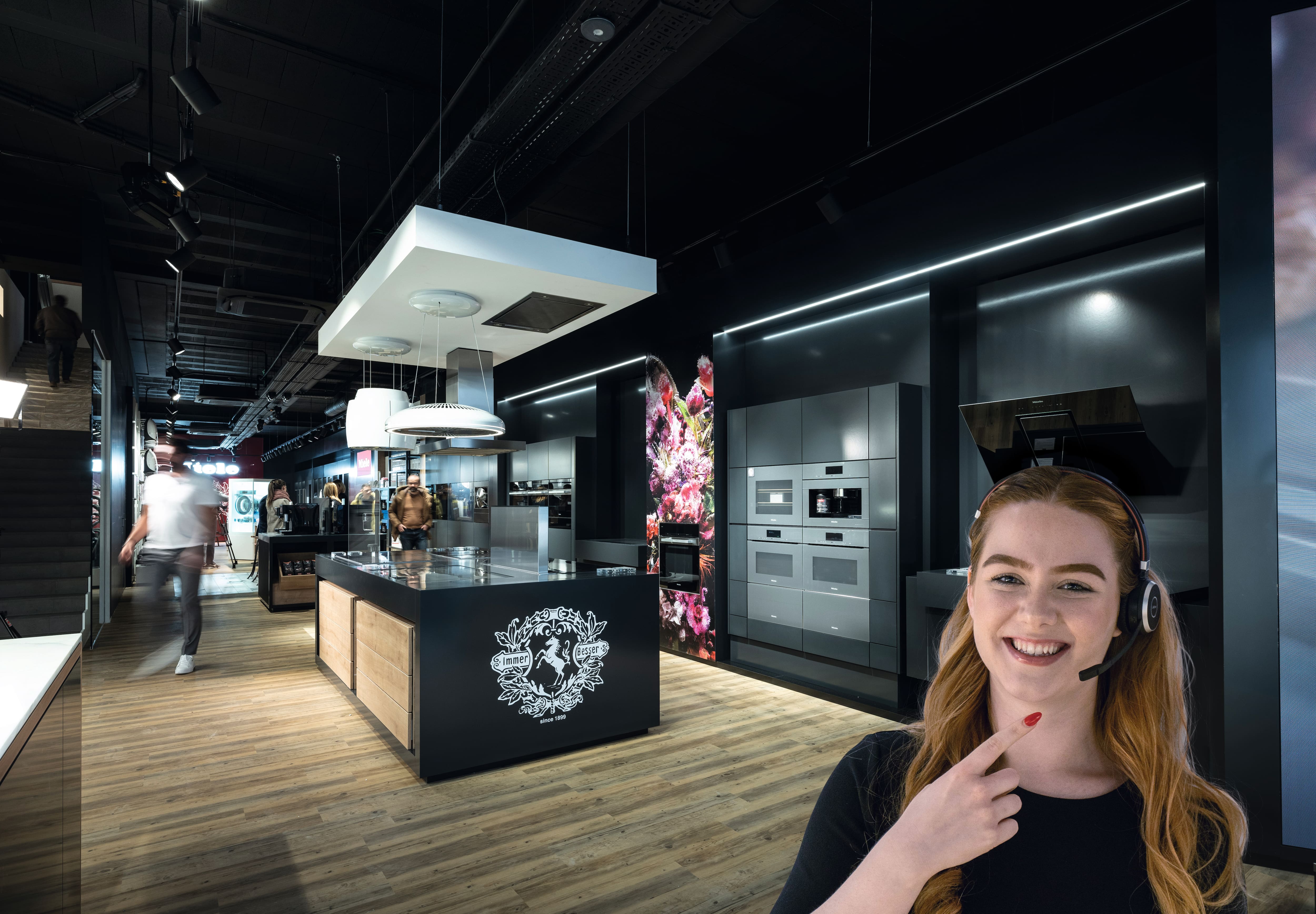 Moderne keukenshowroom met strakke zwarte apparaten, kookeiland, houten vloeren en een medewerker met headset. Moderne keukenshowroom met strakke zwarte apparaten, kookeiland, houten vloeren en een medewerker met headset.
