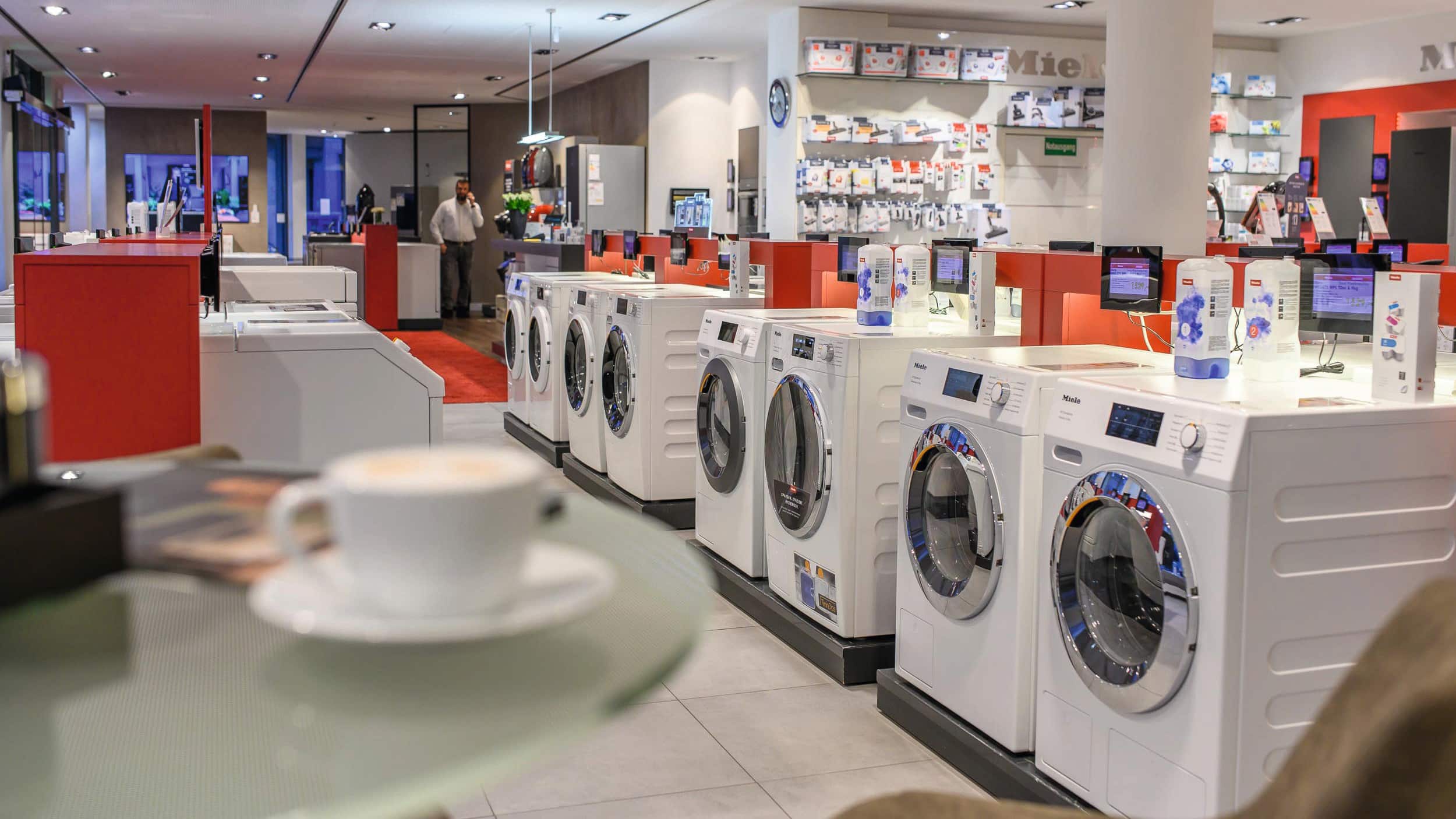 Moderner Gerätegeschäft-Innenraum mit einer Reihe von weißen Frontlader-Waschmaschinen, roten Akzenttheken mit Produktinformationsbildschirmen, Regalen mit Zubehör im Hintergrund und einer Tasse Kaffee im Vordergrund.