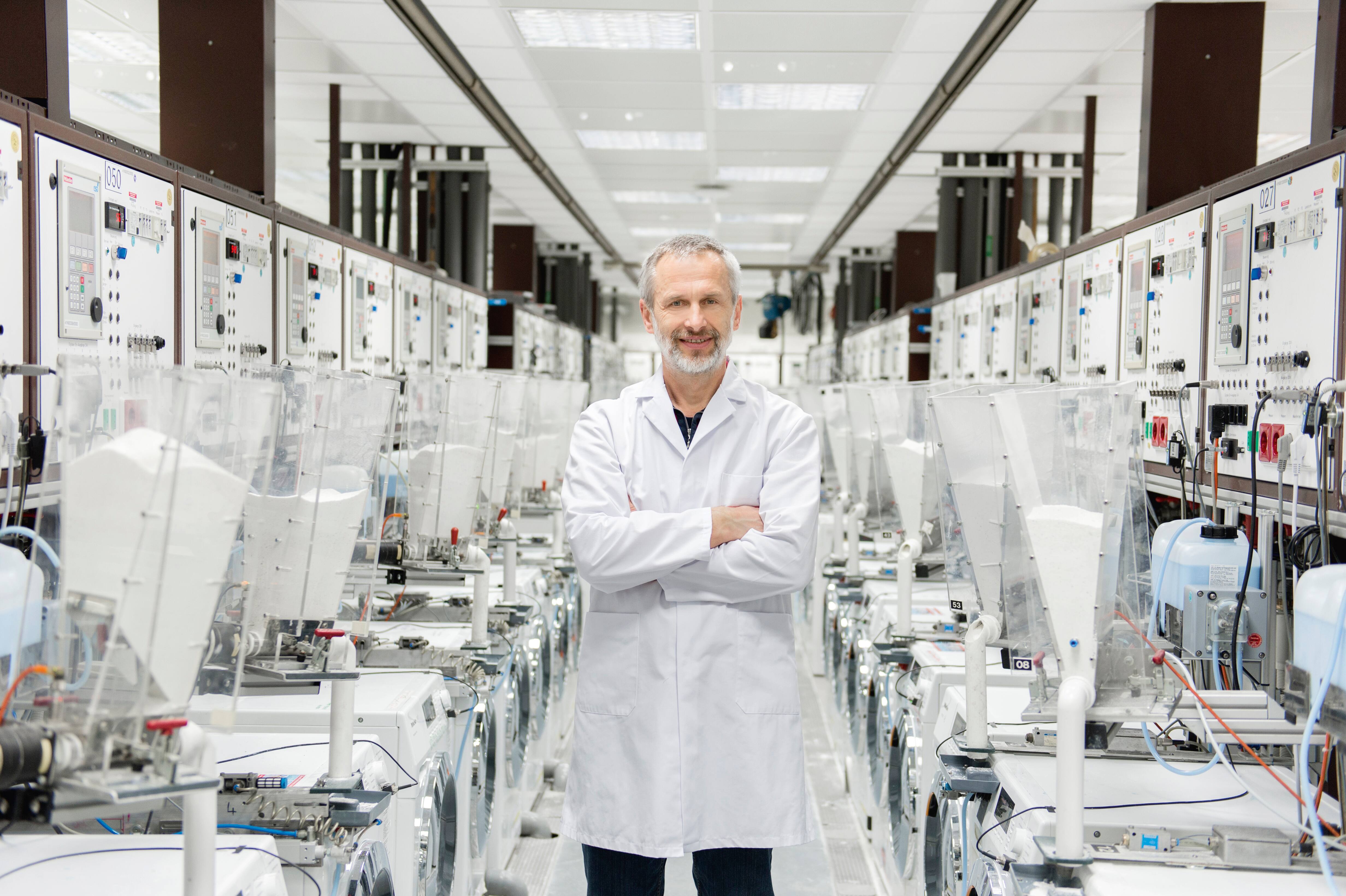 Un ingeniero con bata blanca de laboratorio está de pie, con los brazos cruzados, dentro de un laboratorio de pruebas de electrodomésticos de alta tecnología repleto de máquinas. Un ingeniero con bata blanca de laboratorio está de pie, con los brazos cruzados, dentro de un laboratorio de pruebas de electrodomésticos de alta tecnología repleto de máquinas.