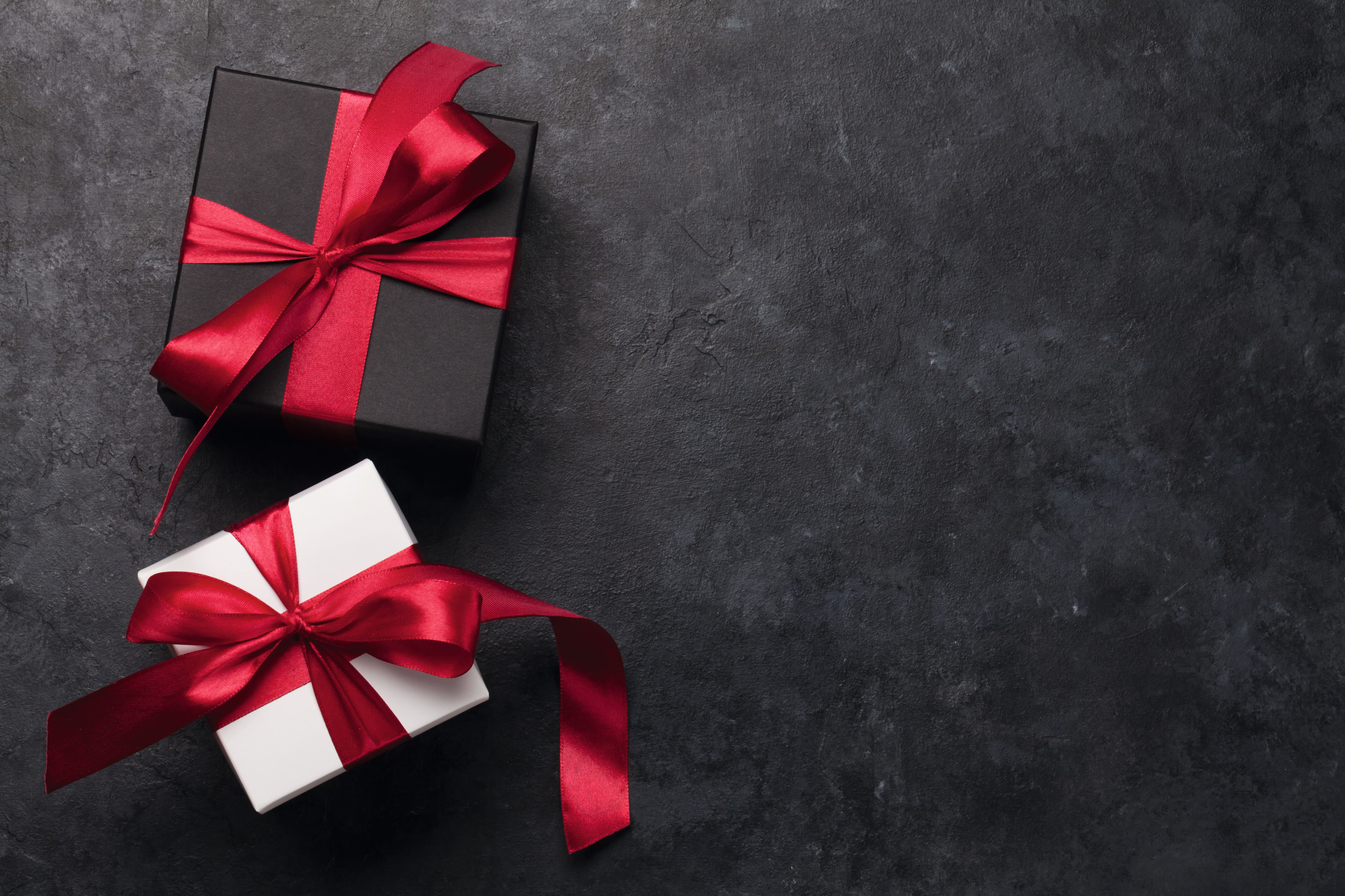 Two gift boxes with red satin ribbons—a black box and a white box—on a dark textured surface with ample copy space on the right. Two gift boxes with red satin ribbons—a black box and a white box—on a dark textured surface with ample copy space on the right.