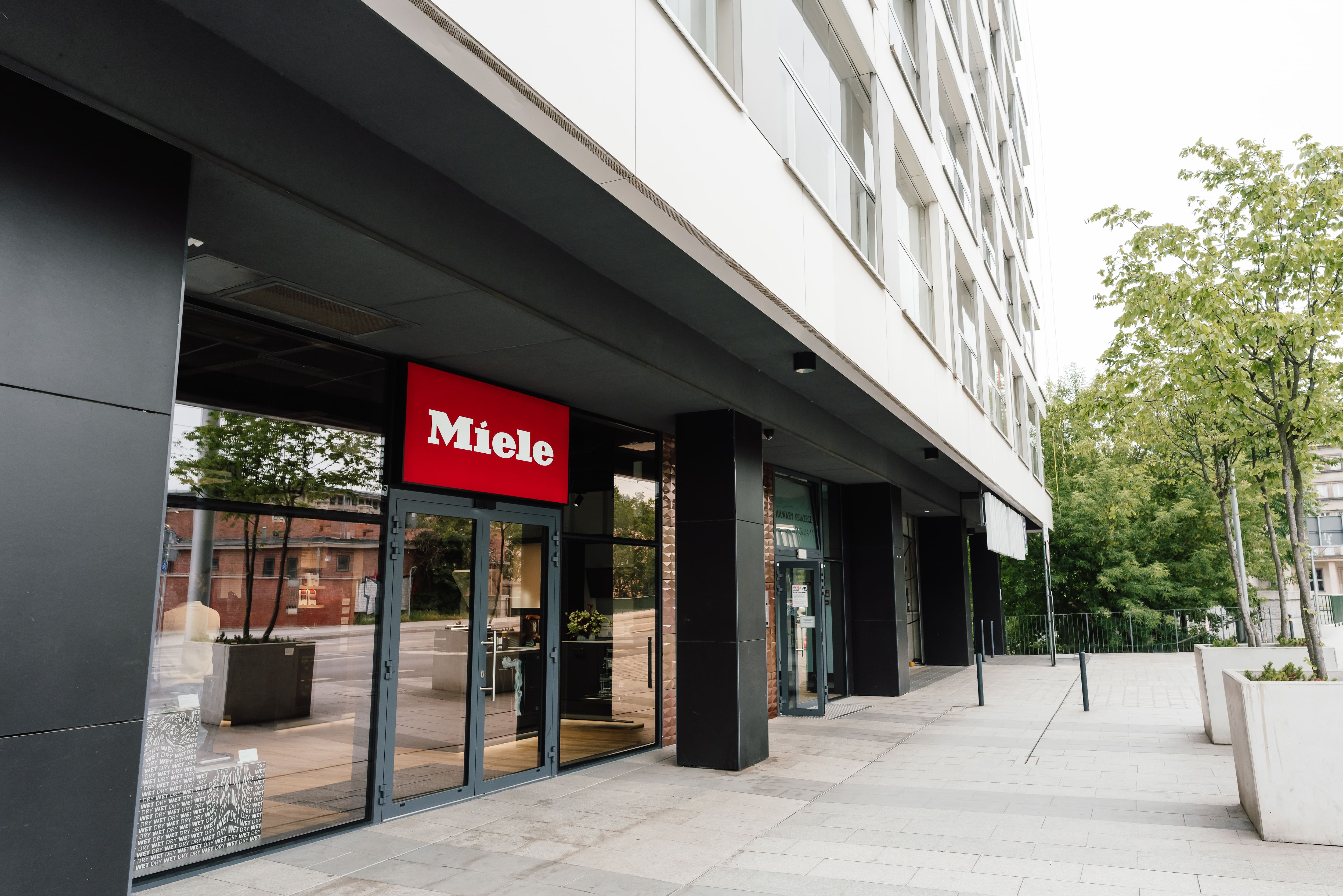 Street-level view of a modern building with a Miele storefront, glass doors, and tree-lined sidewalk.