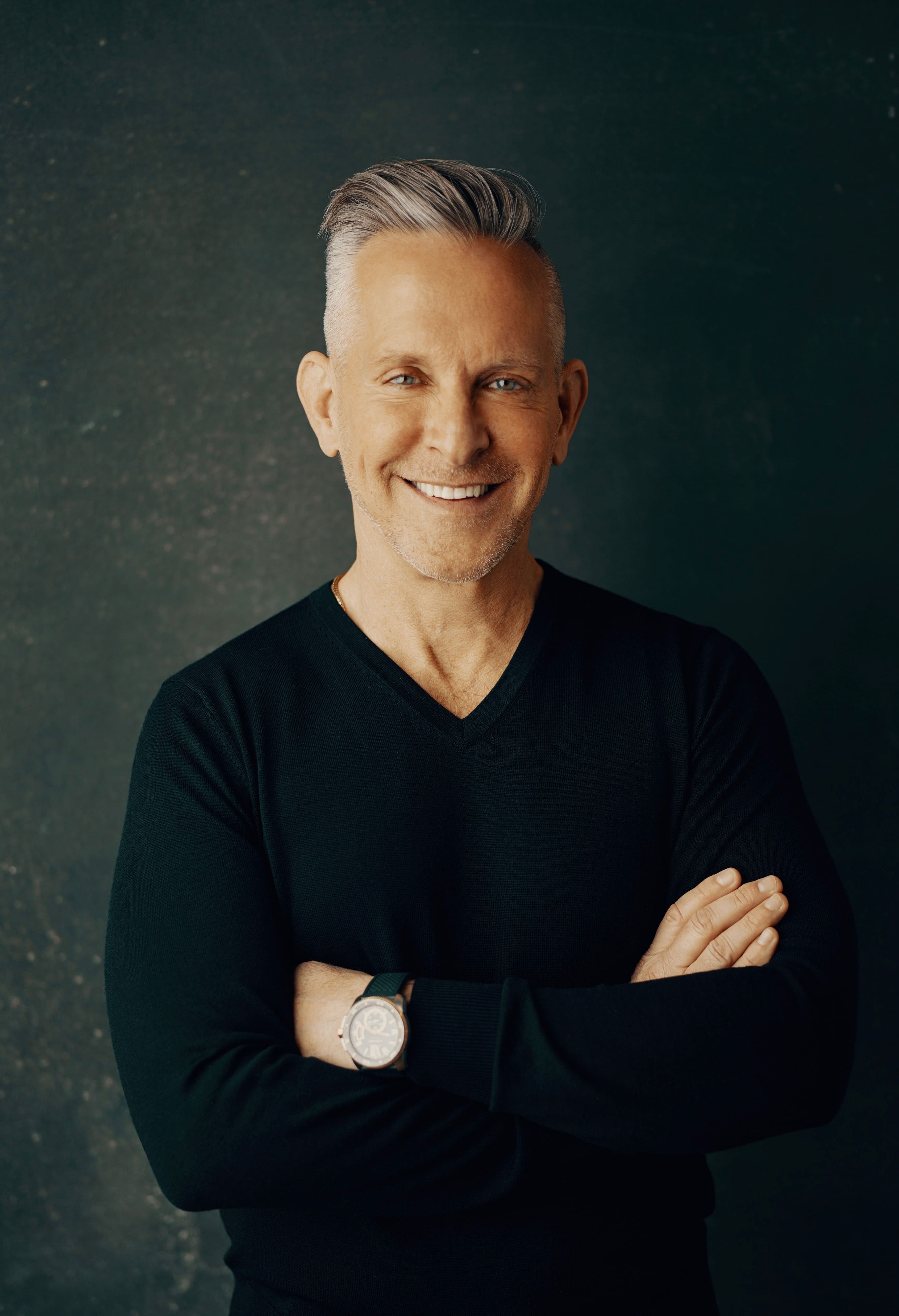 Smiling middle-aged man with gray hair in a black V-neck, arms crossed with a watch, against a dark backdrop.