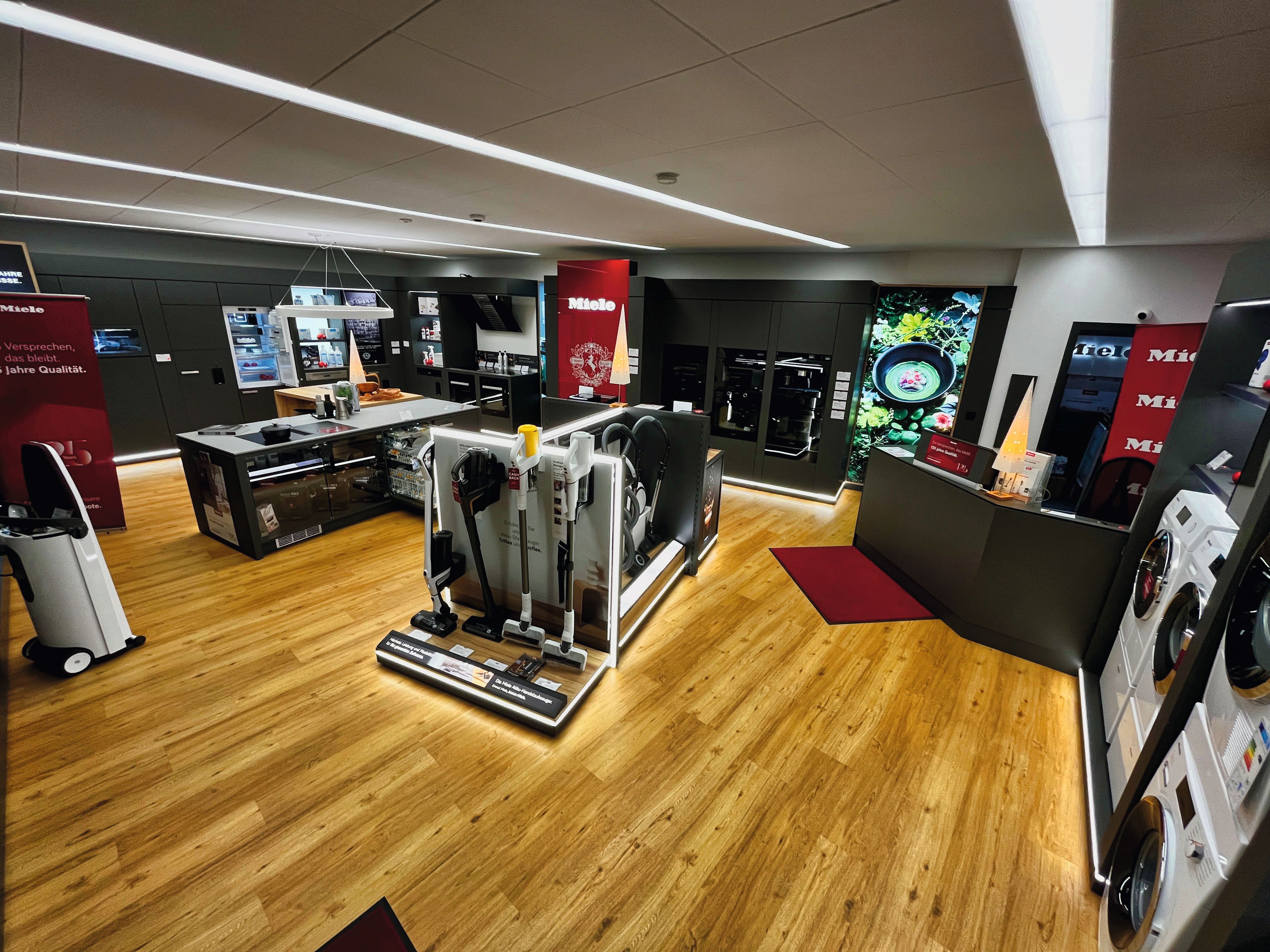 Moderner Geräteschauraum mit Miele-Branding, Holzböden und Ausstellungsinseln mit kabellosen Staubsaugern, Waschmaschinen und Einbauöfen unter eleganter Deckenbeleuchtung.
