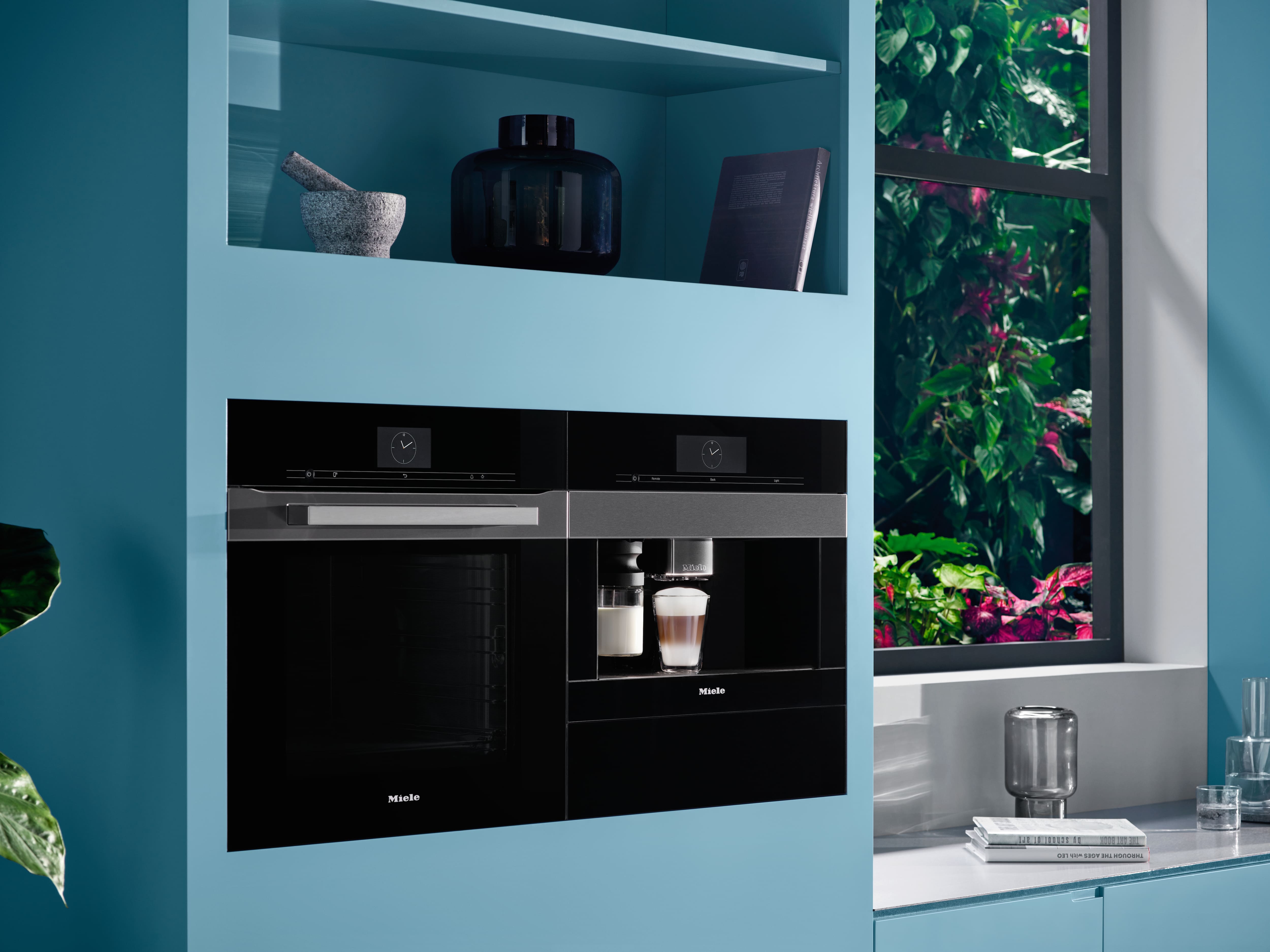 Sleek black built-in oven and coffee machine integrated into blue cabinetry, next to a window with lush greenery and a countertop. Sleek black built-in oven and coffee machine integrated into blue cabinetry, next to a window with lush greenery and a countertop.