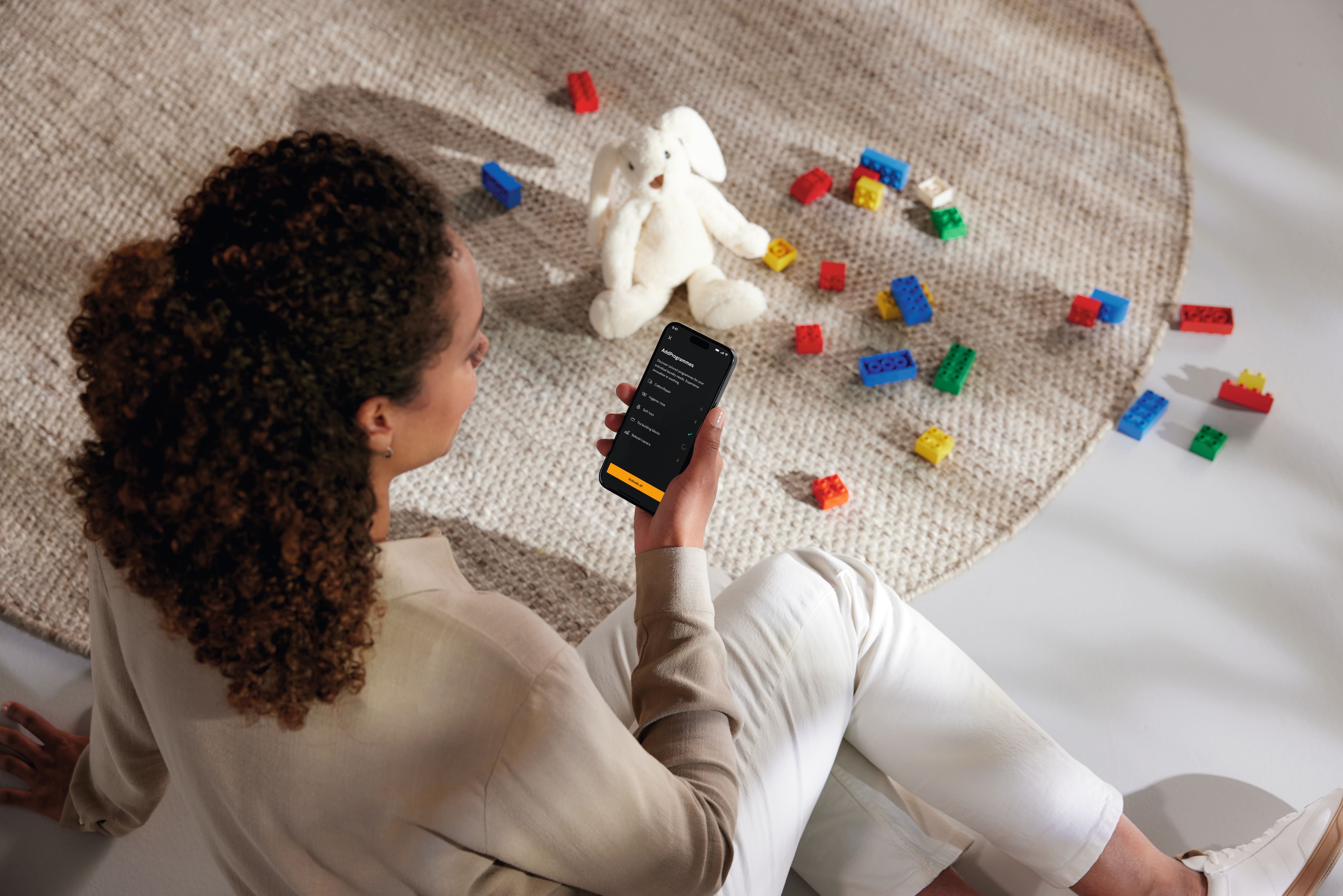 Una persona sentada en el suelo consulta una aplicación del móvil junto a una alfombra redonda en la que hay esparcidos bloques de juguete de colores y un conejito de peluche blanco. Una persona sentada en el suelo consulta una aplicación del móvil junto a una alfombra redonda en la que hay esparcidos bloques de juguete de colores y un conejito de peluche blanco.