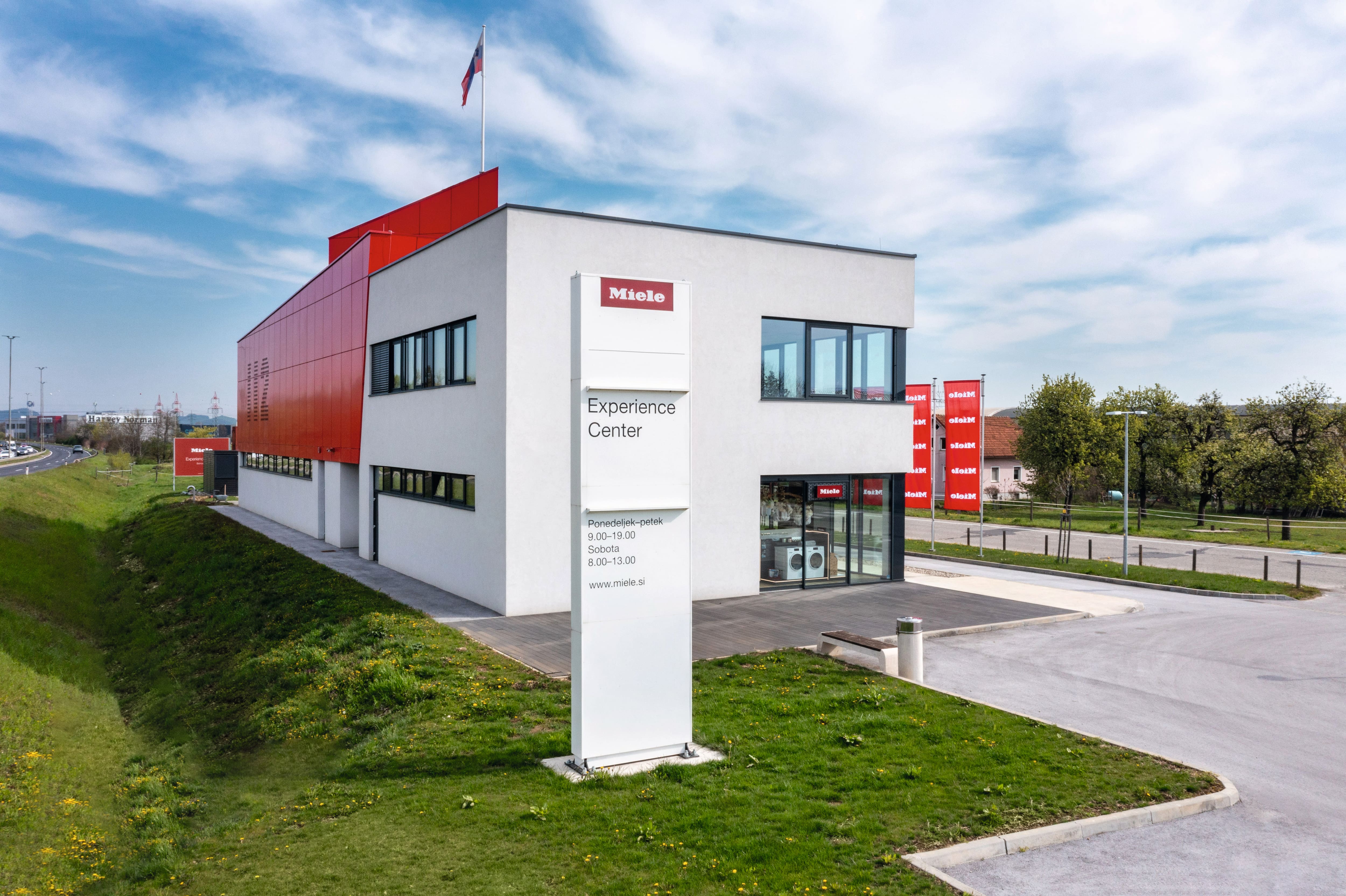 Sodobna bela in rdeča stavba Miele Experience Center z visokim znakom, steklenim vhodom in parkiriščem pod modrim nebom.