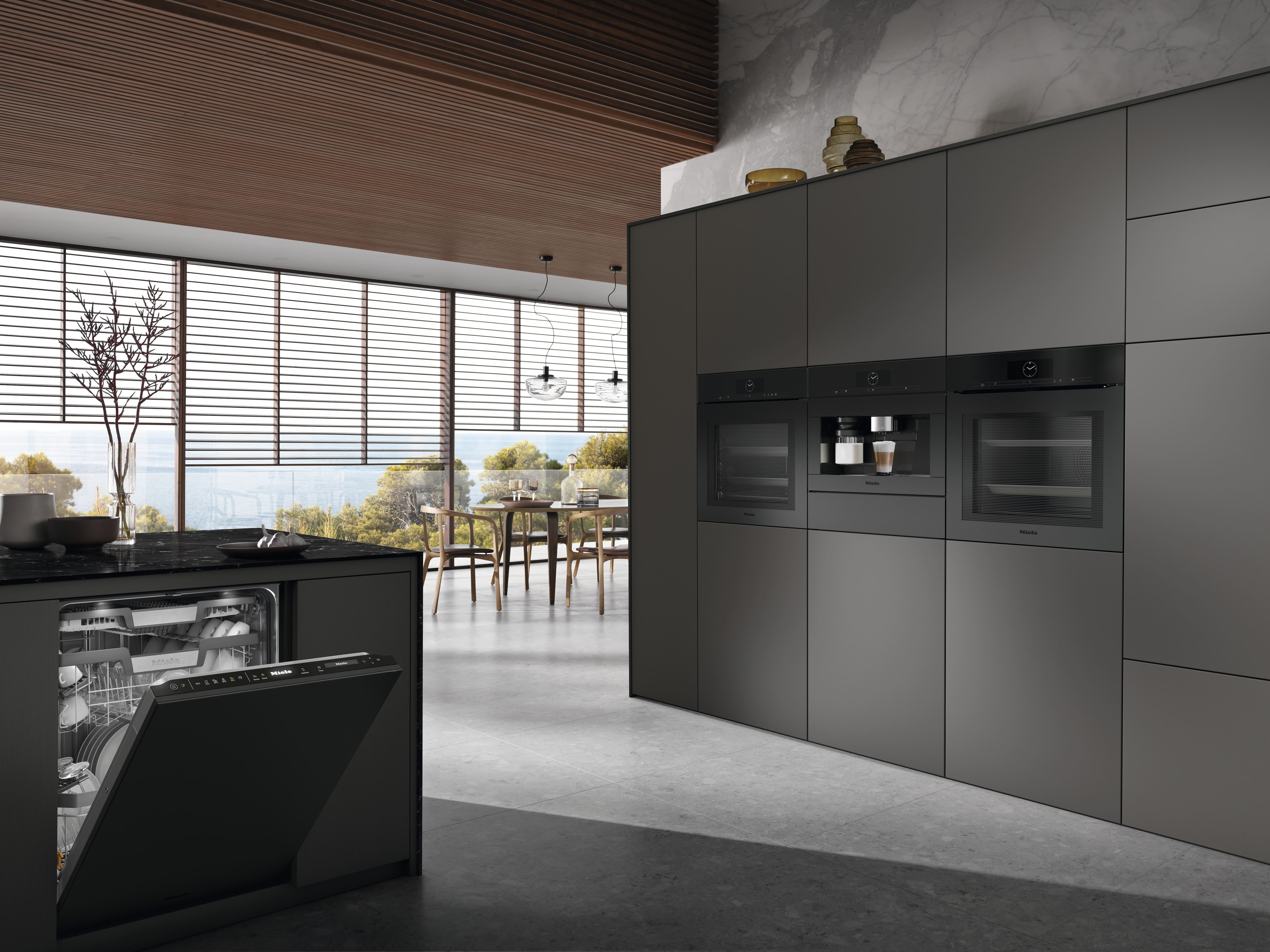 Sleek modern kitchen with Miele built‑in wall ovens and a Miele coffee machine, an open Miele dishwasher in a dark island, marble backsplash, floor‑to‑ceiling windows with blinds, and a dining area overlooking trees and water. Sleek modern kitchen with Miele built‑in wall ovens and a Miele coffee machine, an open Miele dishwasher in a dark island, marble backsplash, floor‑to‑ceiling windows with blinds, and a dining area overlooking trees and water.