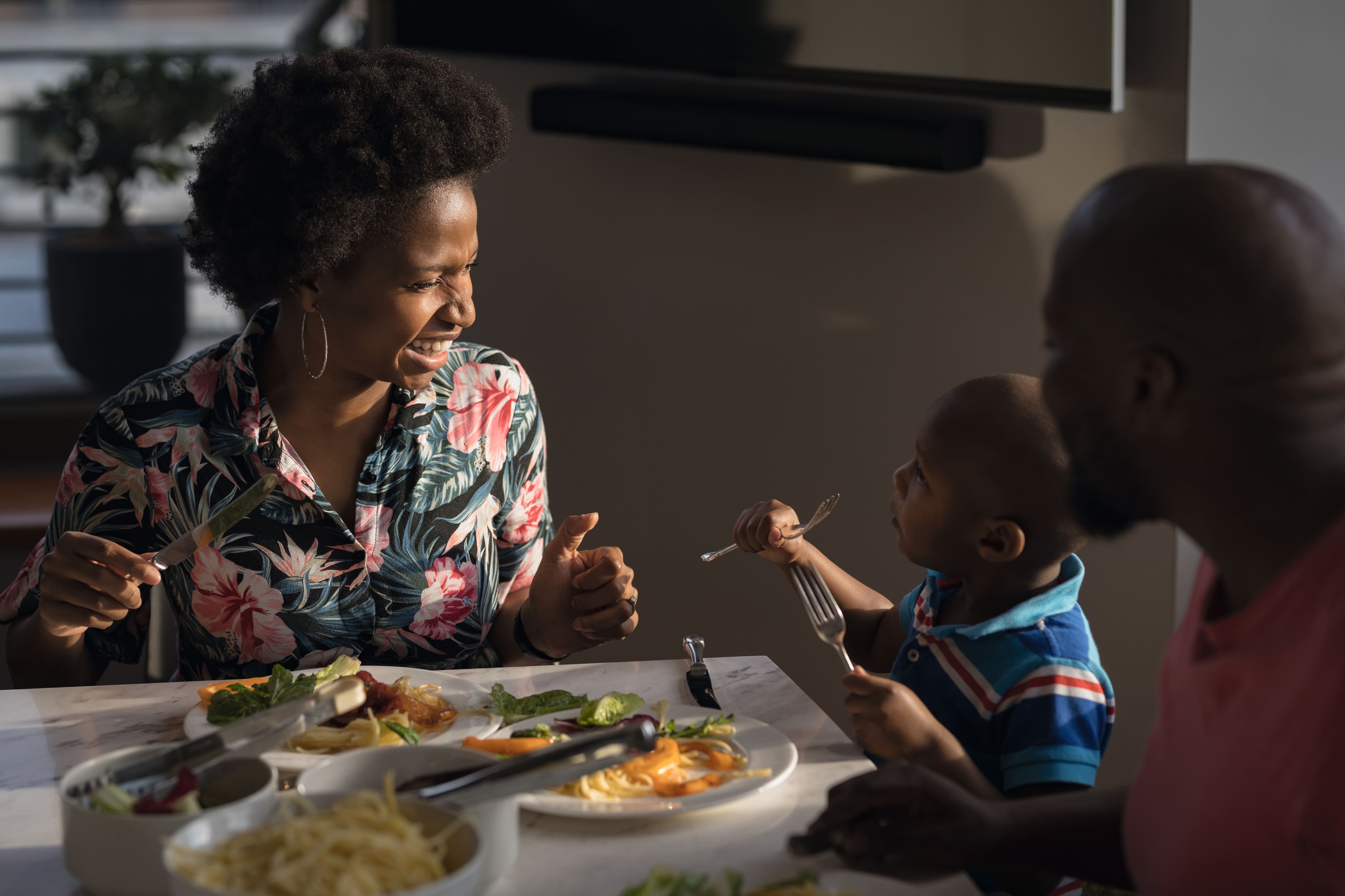Famiglia sorridente condivide una cena informale a casa mentre un bambino tiene le posate, con piatti di pasta e insalata. Famiglia sorridente condivide una cena informale a casa mentre un bambino tiene le posate, con piatti di pasta e insalata.