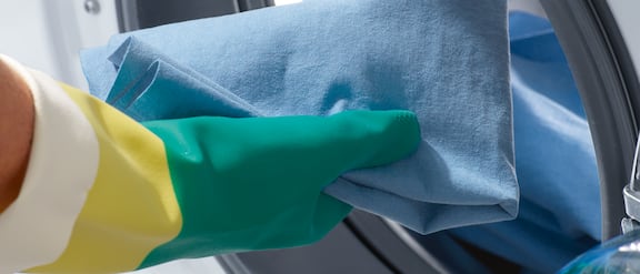 Händer med gummihandskar lägger blåa städdukar i en professionell tvättmaskin.