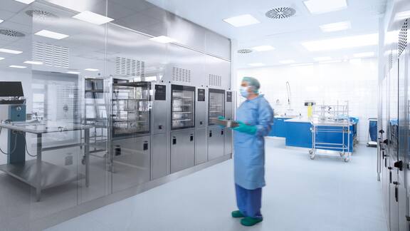 Pracovník v nemocničním oblečení stojí před řadou šedých sterilizačních přístrojů.