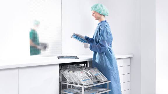Egy sebészköpenyt viselő betegápoló betölt egy Miele Professional asztali tisztító- és fertőtlenítő készüléket.